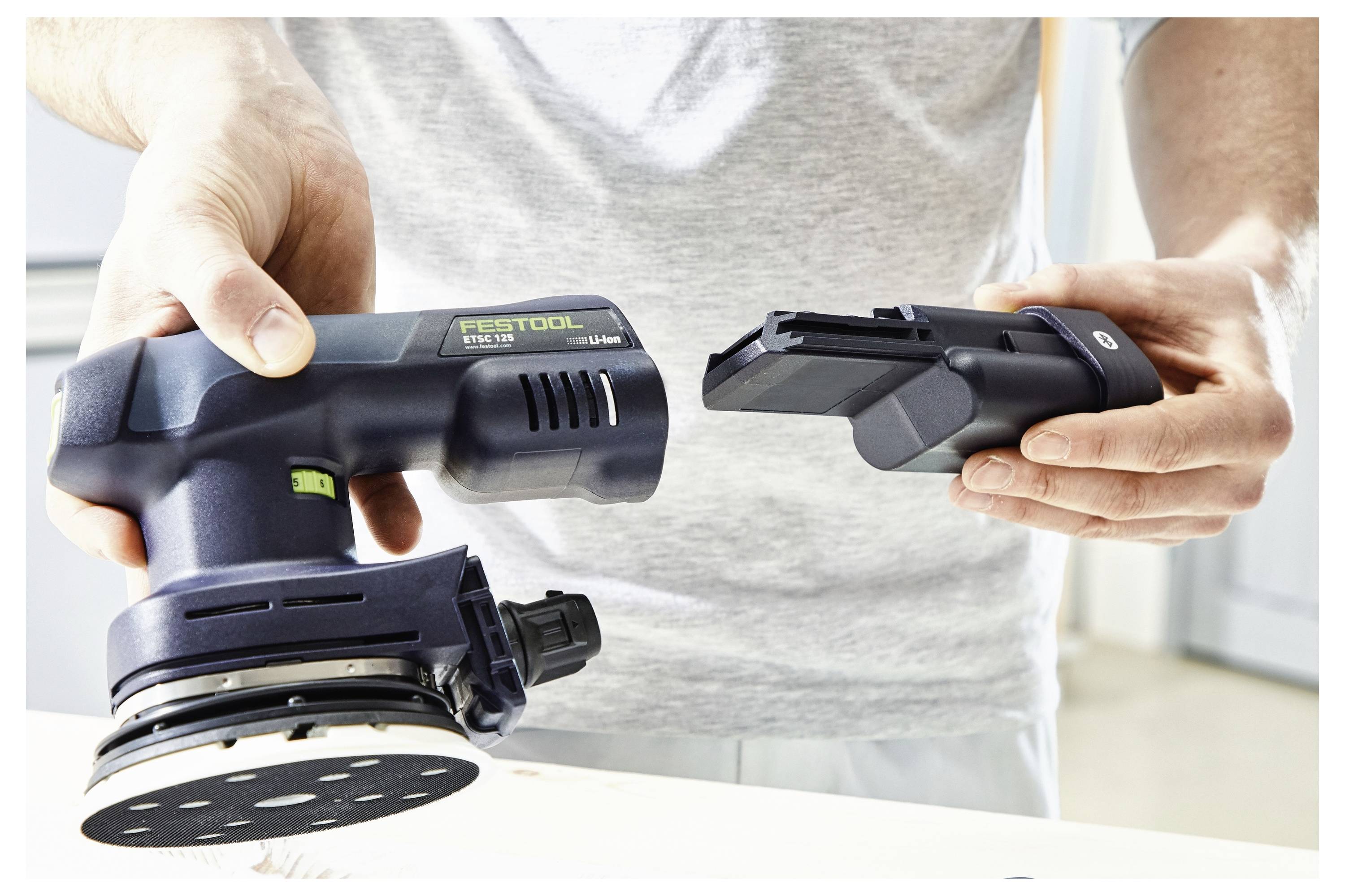 En person som håller en svart handhållen slipmaskin i ena handen och ett avtakbart batteri i den andra, vilket antyder att verktyget är batteridrivet.