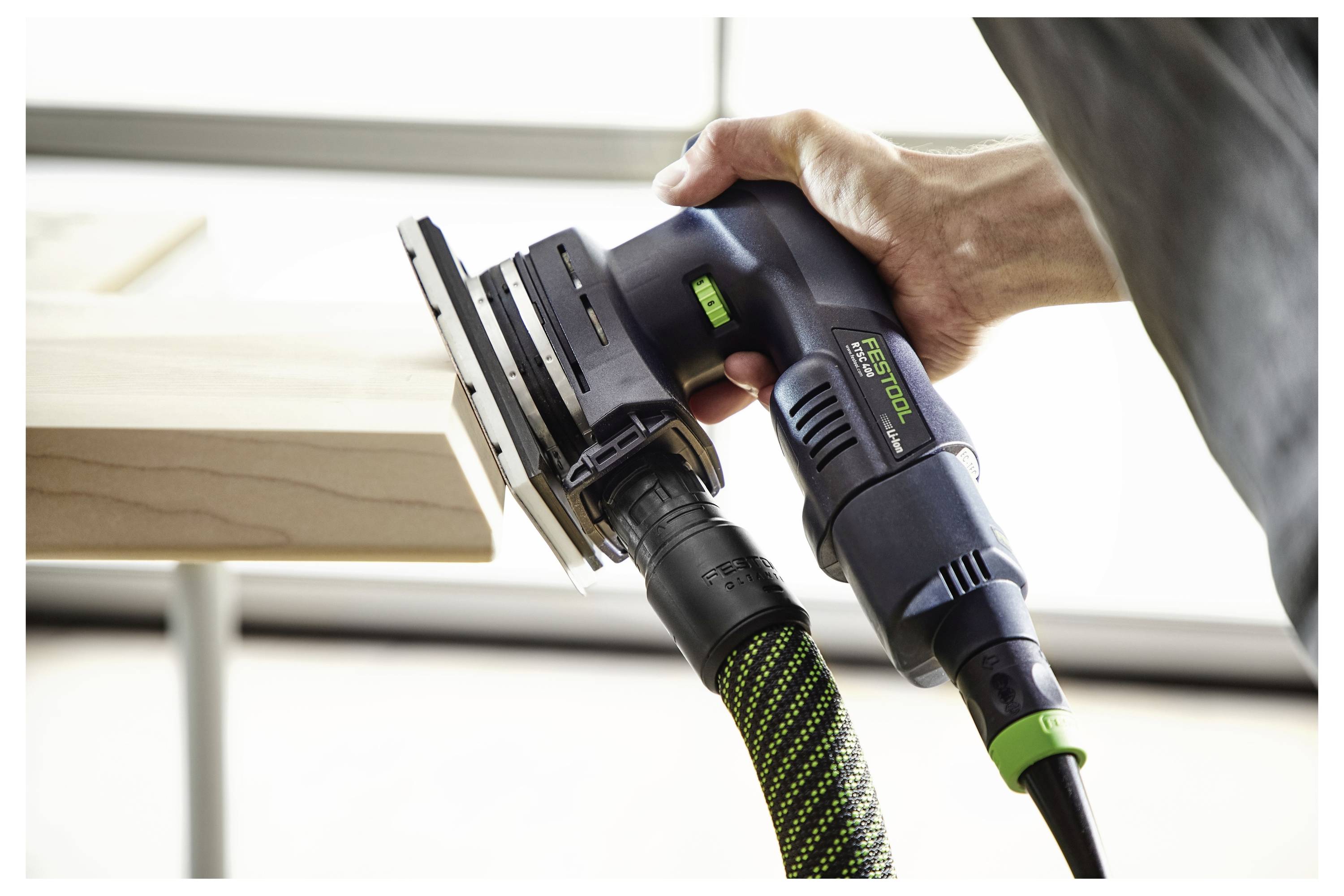 Närbild av en persons hand som använder en handhållen elektrisk slipmaskin på en träyta, vilket visar fokuserad slipningsåtgärd.