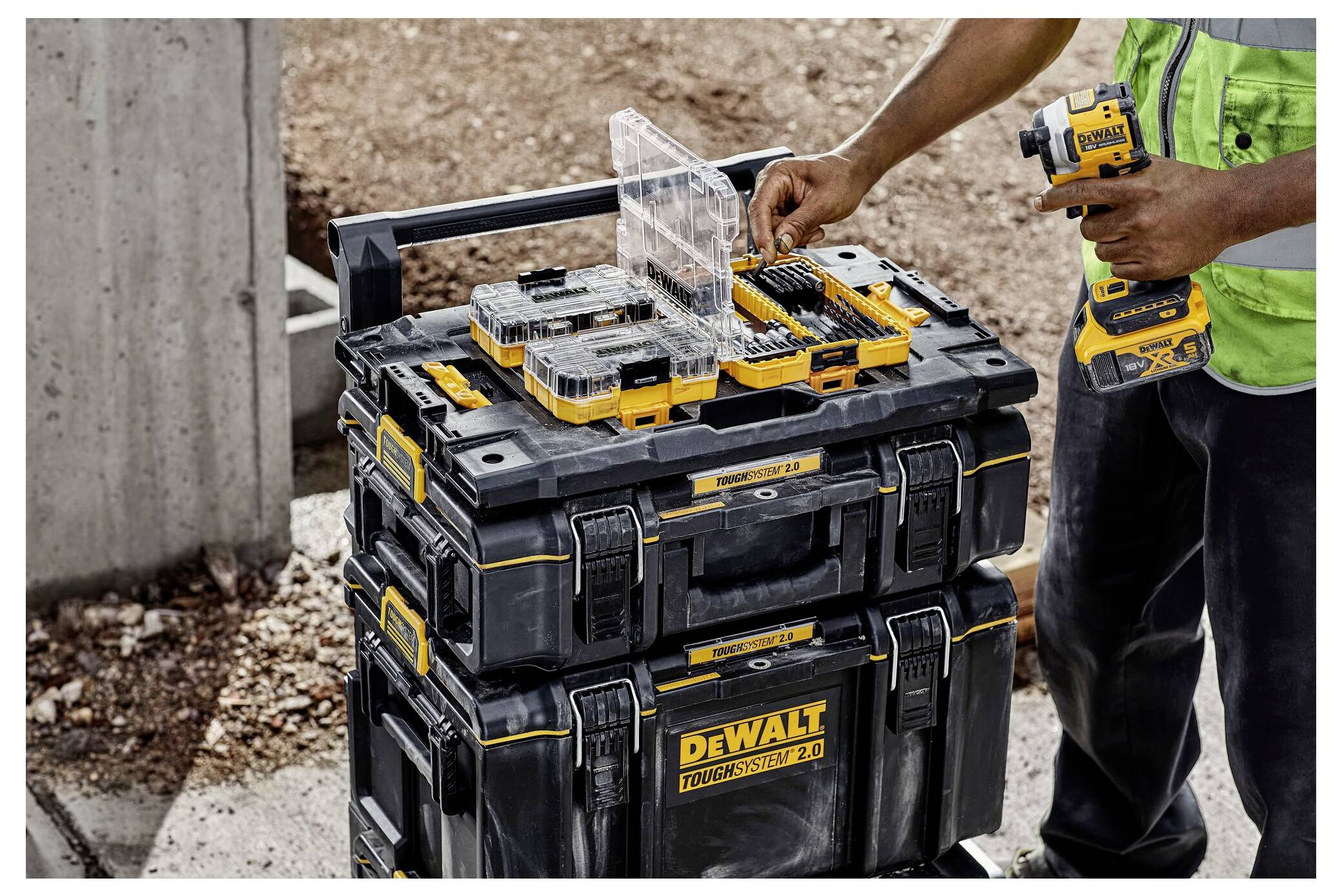 En arbetare arrangerar verktyg i ett svart och gult DEWALT-förvaringsfodral, medan han håller en elborr. Miljön verkar vara på en byggarbetsplats.