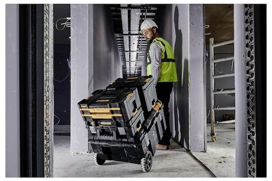 En byggnadsarbetare i en säkerhetshjalm och en säkerhetsvast drar ett staplade verktygsfodral på hjul genom en smal korridor på en byggarbetsplats.