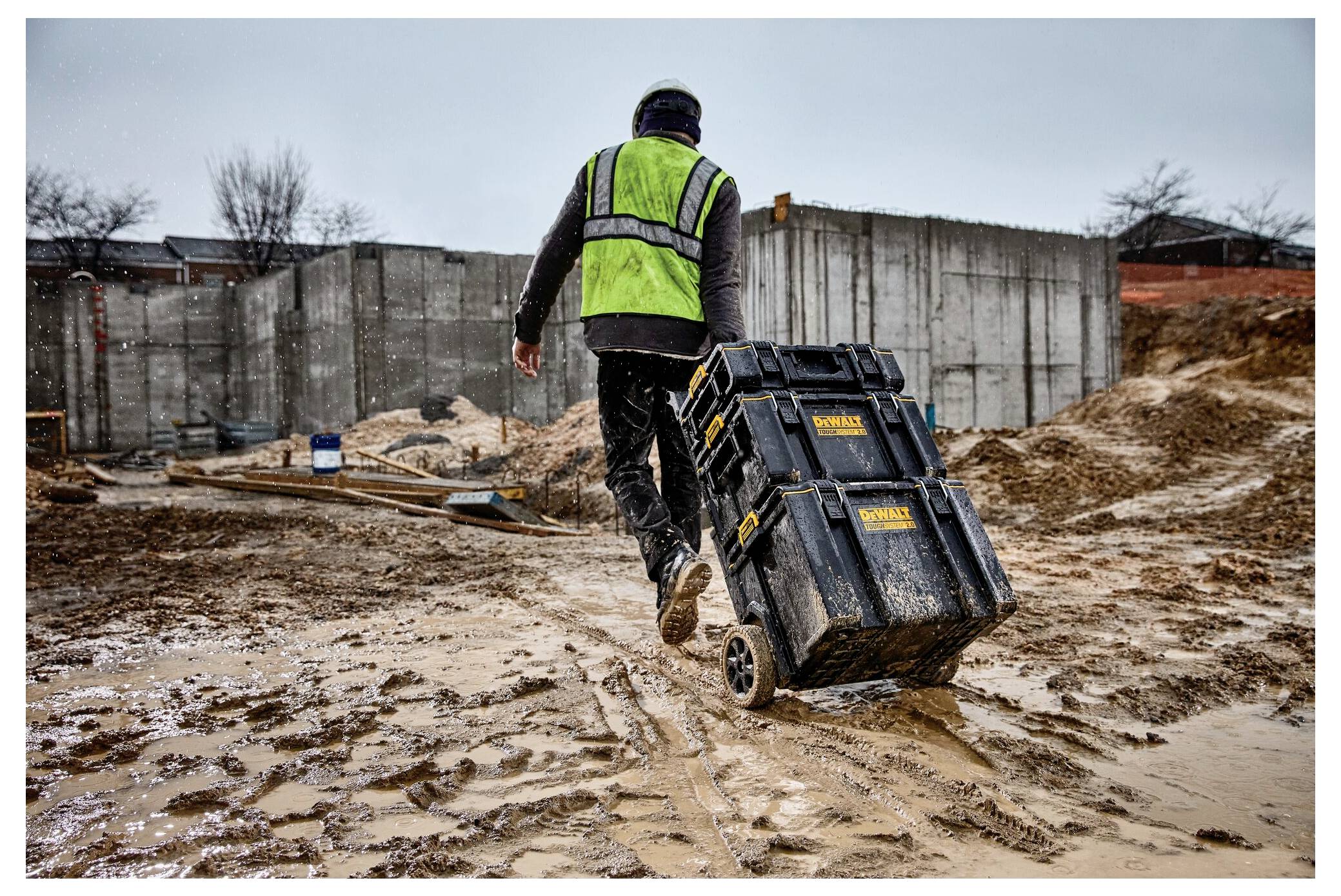 En person i en grön väst och hjälm drar en verktygsvagn över en lerig byggarbetsplats med betongväggar och utspridda material.