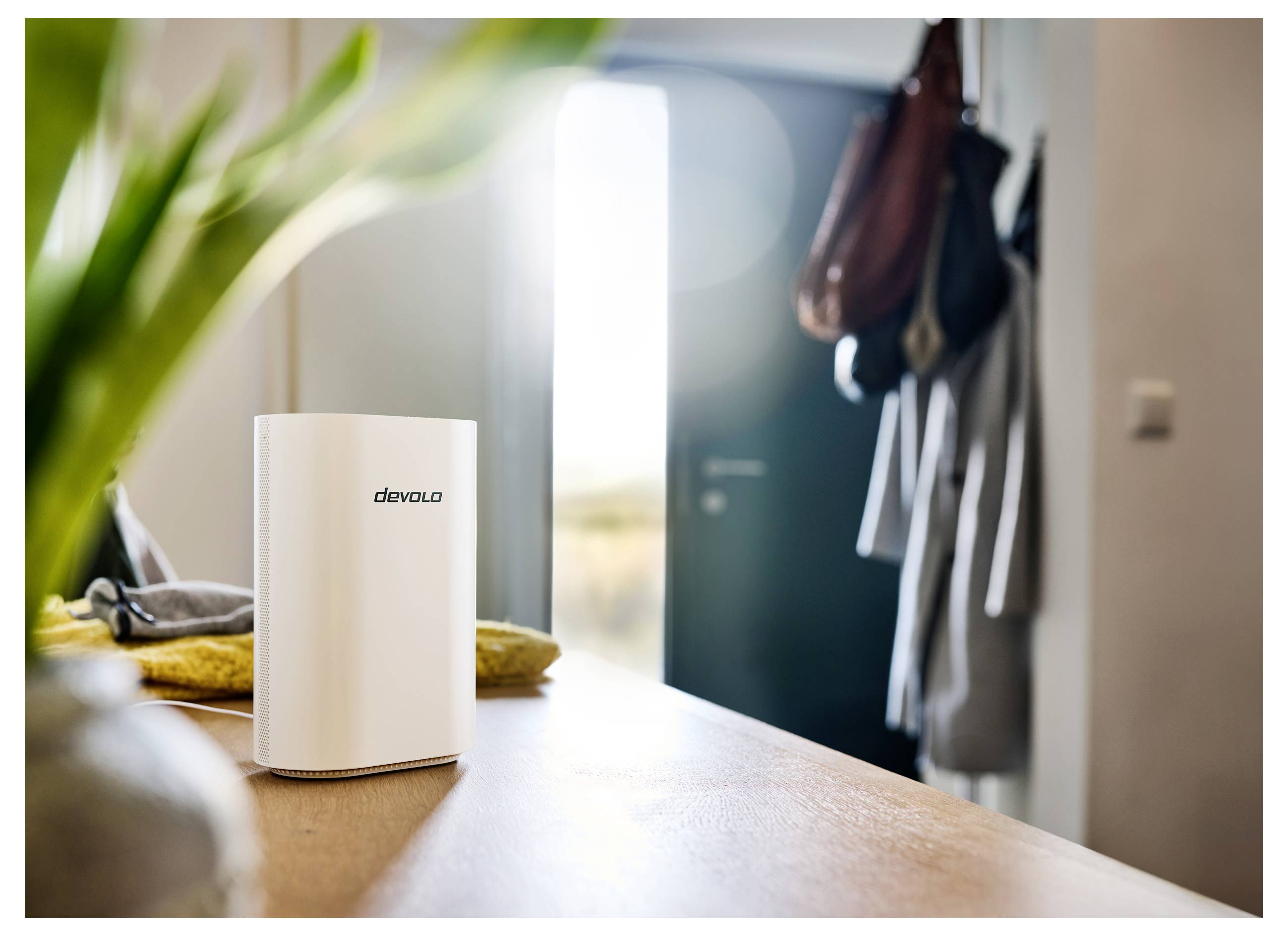En vit elektronisk enhet med "devolo"-märke står på ett trägolv på ett trägolv i ett ljust rum. Bakgrunden visar en öppen dörr och hängande rockar.