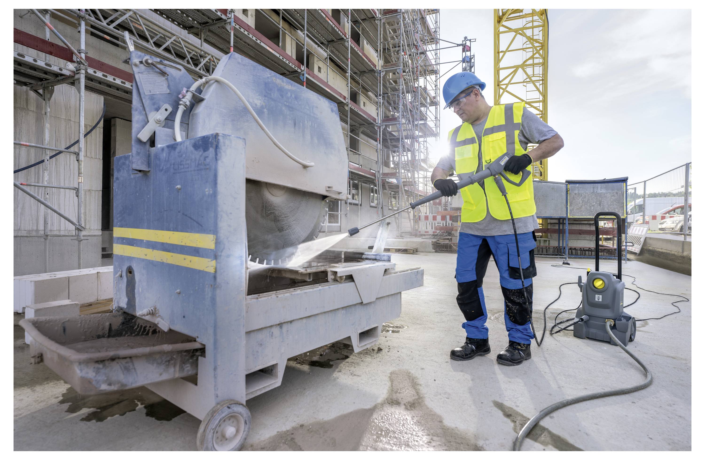 En byggnadsarbetare i skyddsutrustning tvätta en cementblandare med högtryckstvätt på en byggarbetsplats, med ställningar och en gul kran i bakgrunden.
