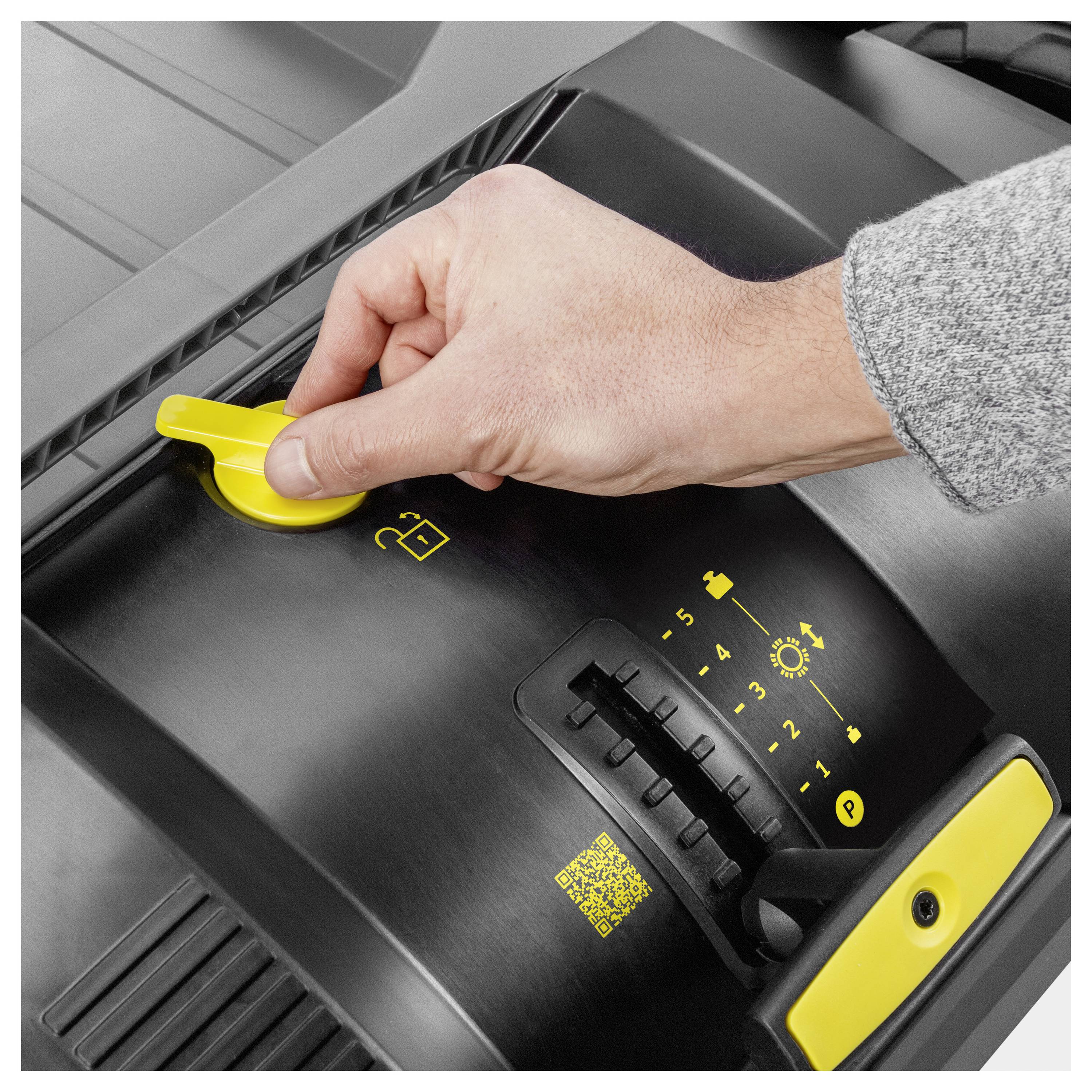 En persons hand justerar en gul spak på en maskin med en numrerad skala och små ikoner, vilket indikerar anpassningsbara inställningar.