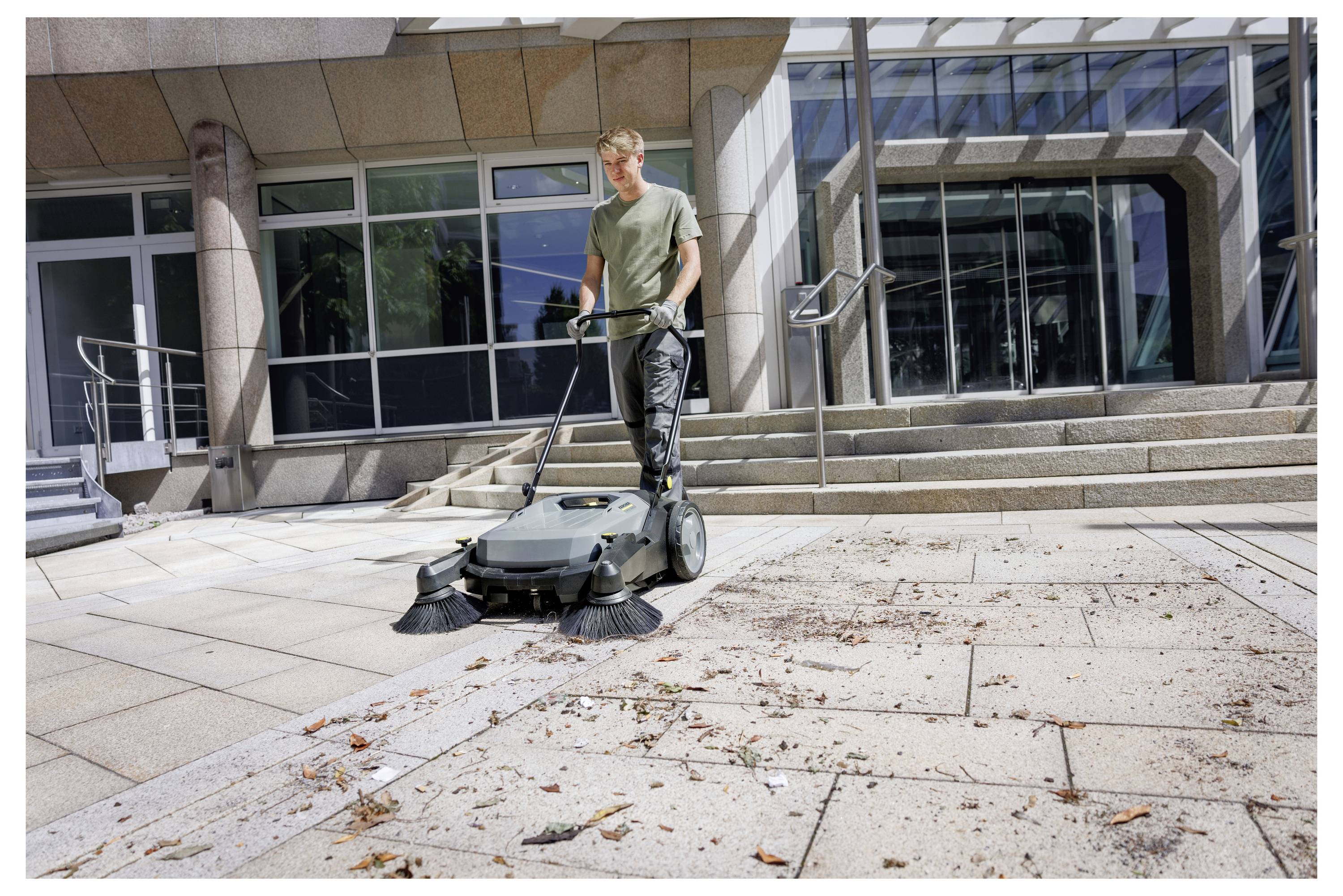 En person använder en utomhuskväst för att städa upp skräp på en stor, kaklad plats utanför en modern byggnad med glas- och metallarkitektur.