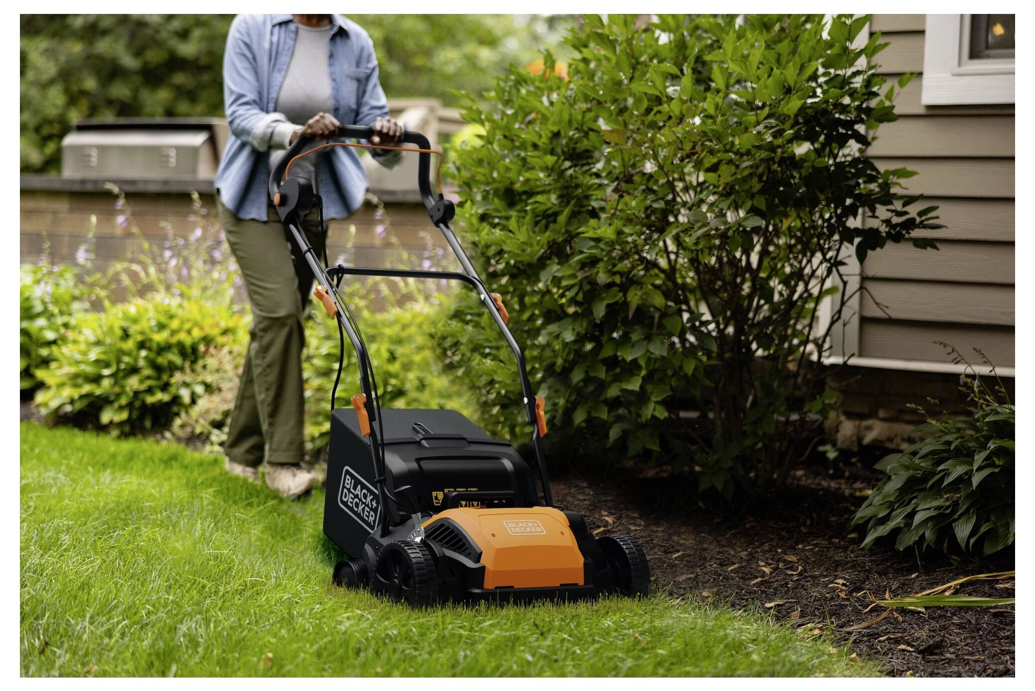 En person klipper gräsmattan med en orange och svart elektrisk gräsklippare nära en planteringsbädd, intill en husfasad.