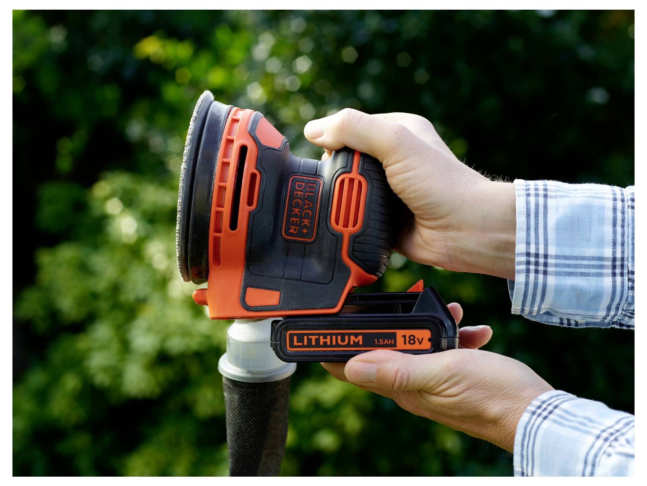 En person som håller en trådlös Black & Decker-slipmaskin, sätter in ett 18V lithiumbatteri, med en trädgårdsbakgrund.