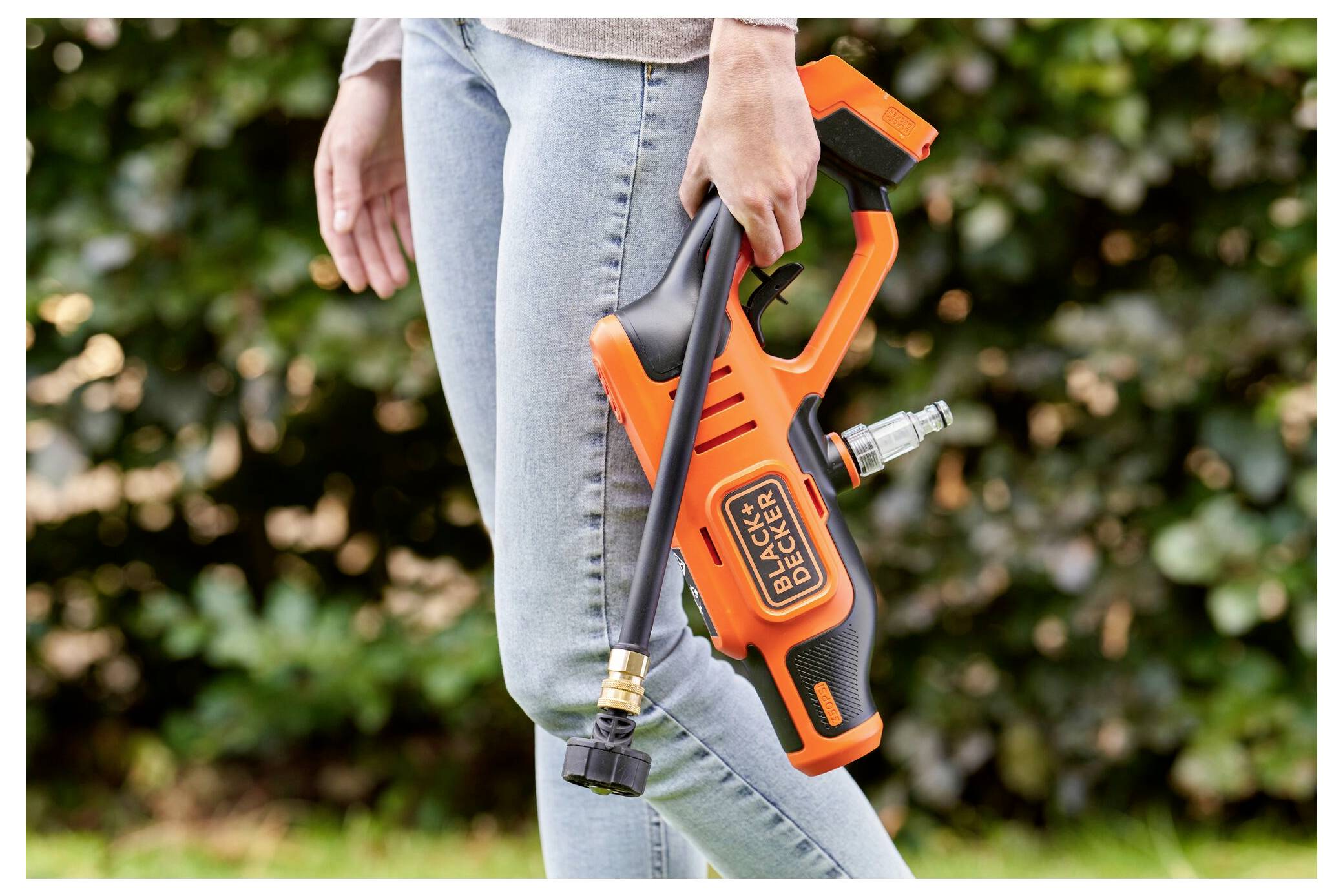 En person som håller en orange Black & Decker högtryckstvätt och står utomhus. Enheten har en lång munstycke som är lämplig för rengöringsuppgifter.