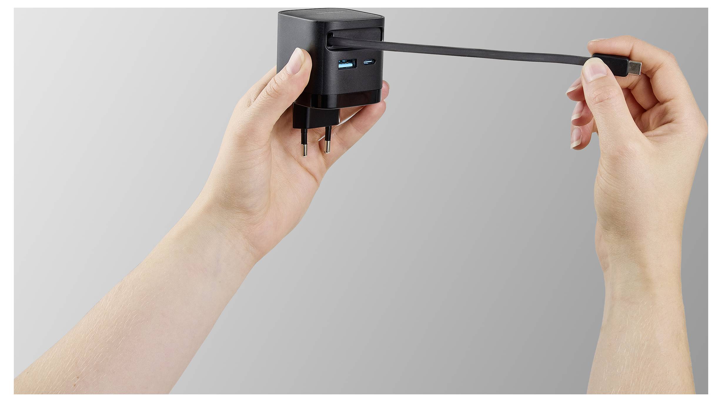 En person demonstrerar den infällbara stickkontakten på en kompakt svart strömadapter med USB-portar, mot en enkel grå bakgrund.