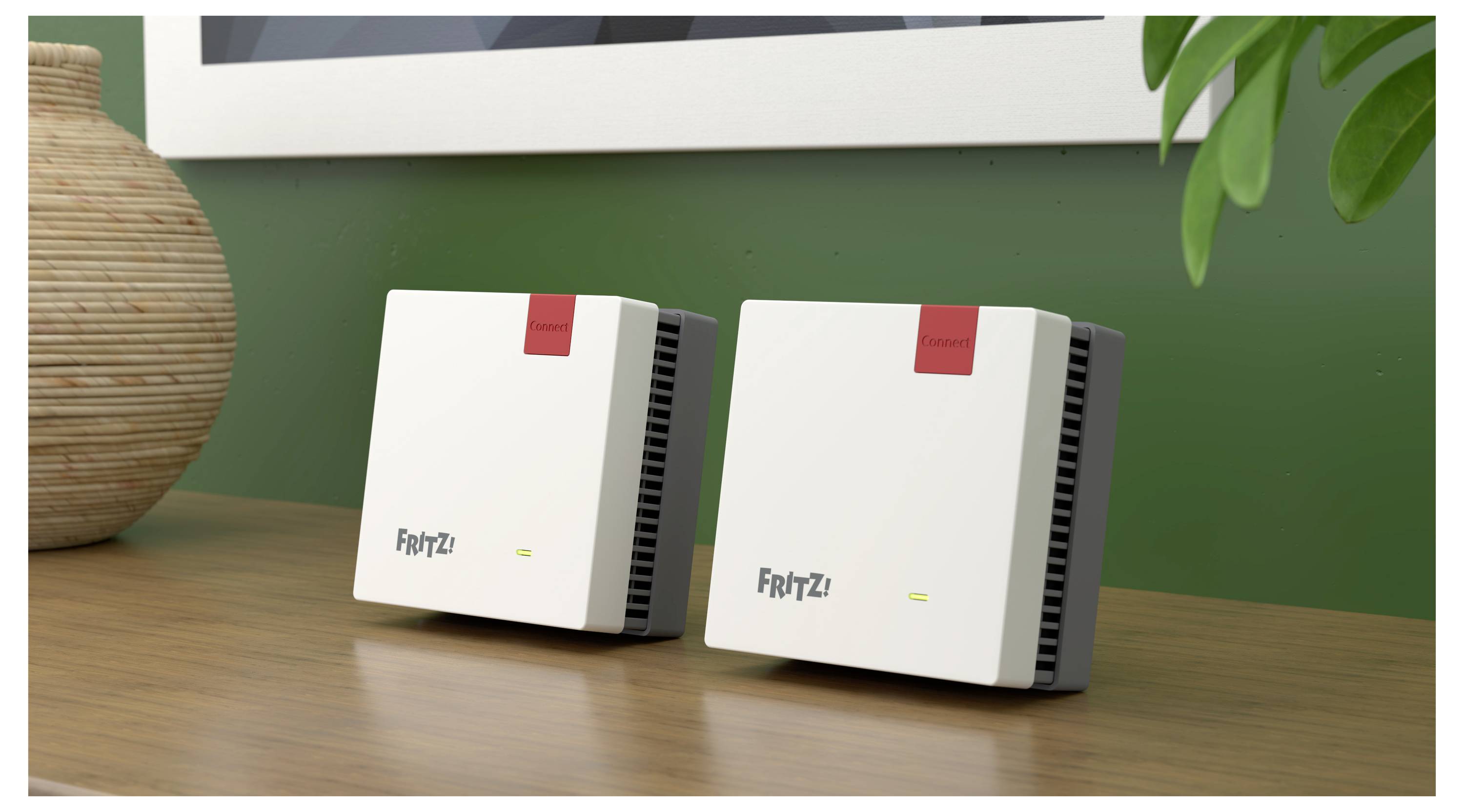 Två vita elektroniska enheter märkta "FRITZ!" är placerade på ett träbord, bredvid en vas. De är små och rektangulära, med ventilerade sidor.