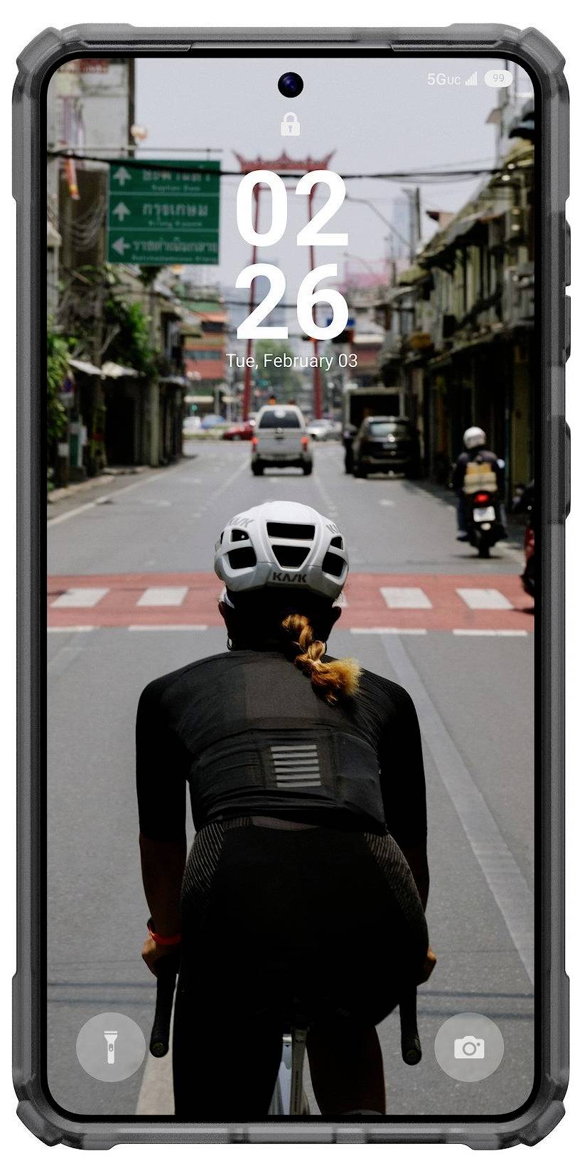En person i cyklutrustning cyklar på en stadsgata omgiven av byggnader, sett på en smartphoneskärm som visar tid och datum.