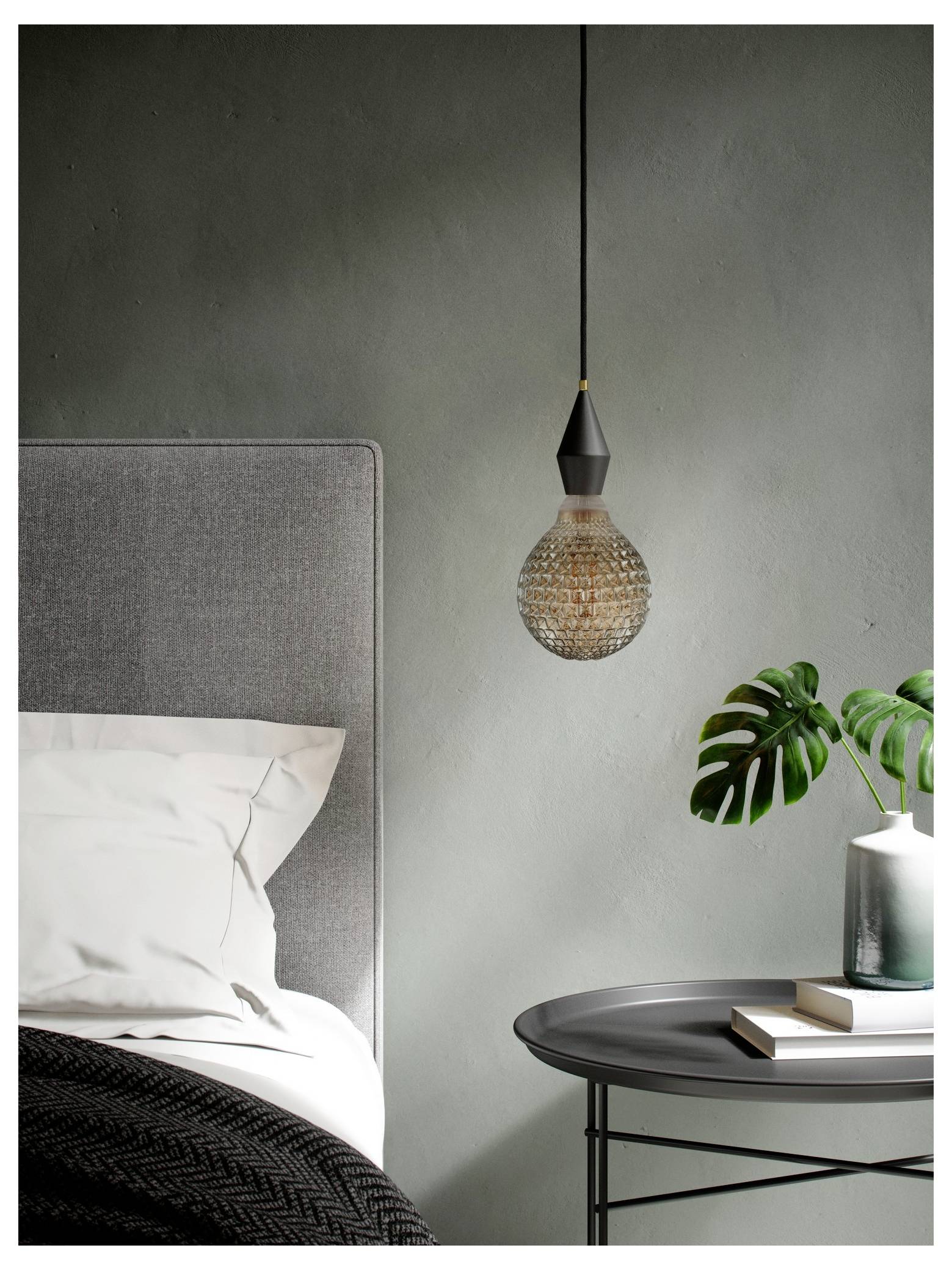 Ett gråt sovrum med en modern hängande lampa, en säng med vita lakan och ett rundt bord som håller böcker och en krukväxt.