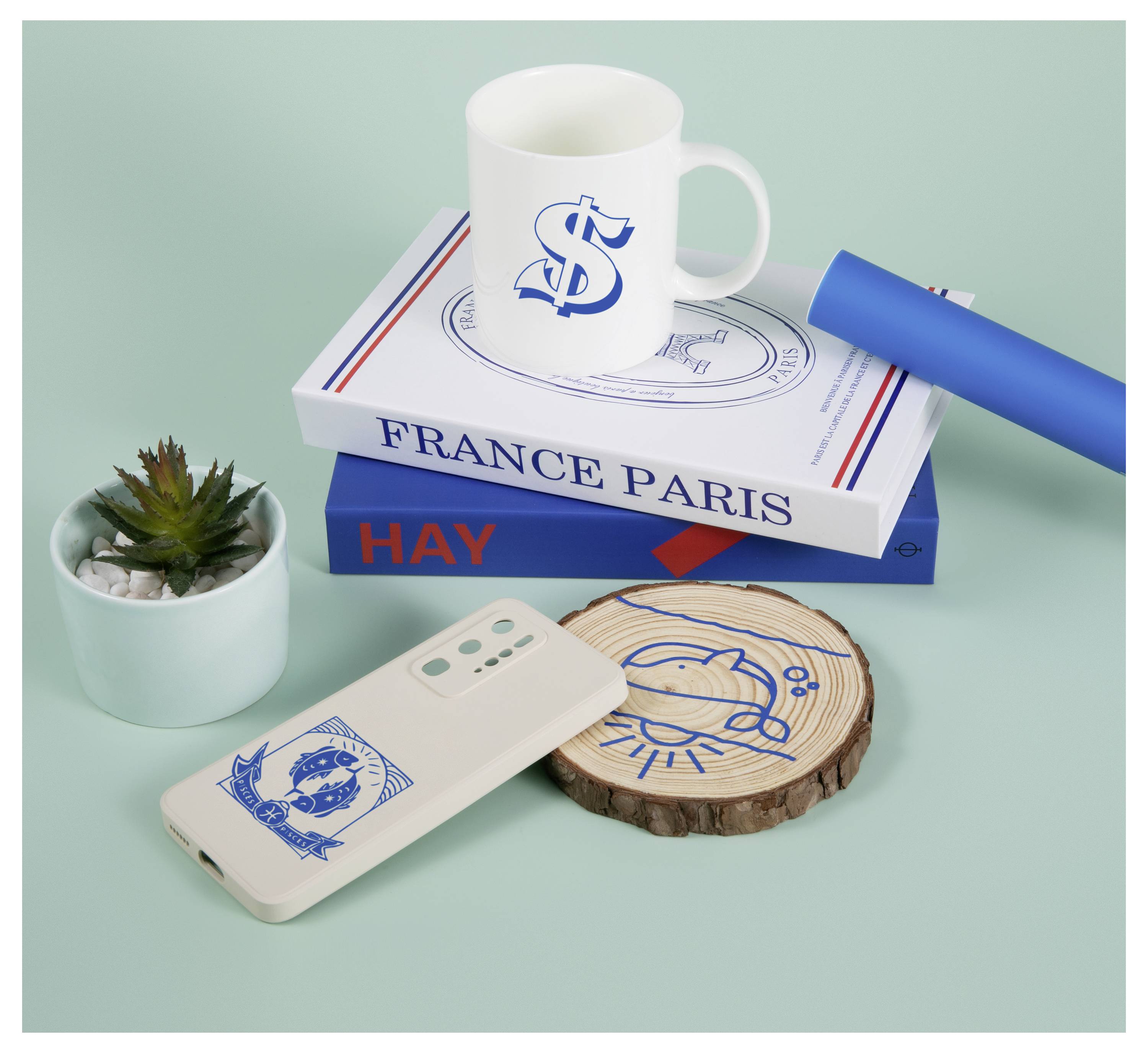 En mugg med en dollartteckendesign, en smartphone, en träunderlägg, en bok med titeln "FRANCE PARIS" och en liten växt på en turkosblå yta.