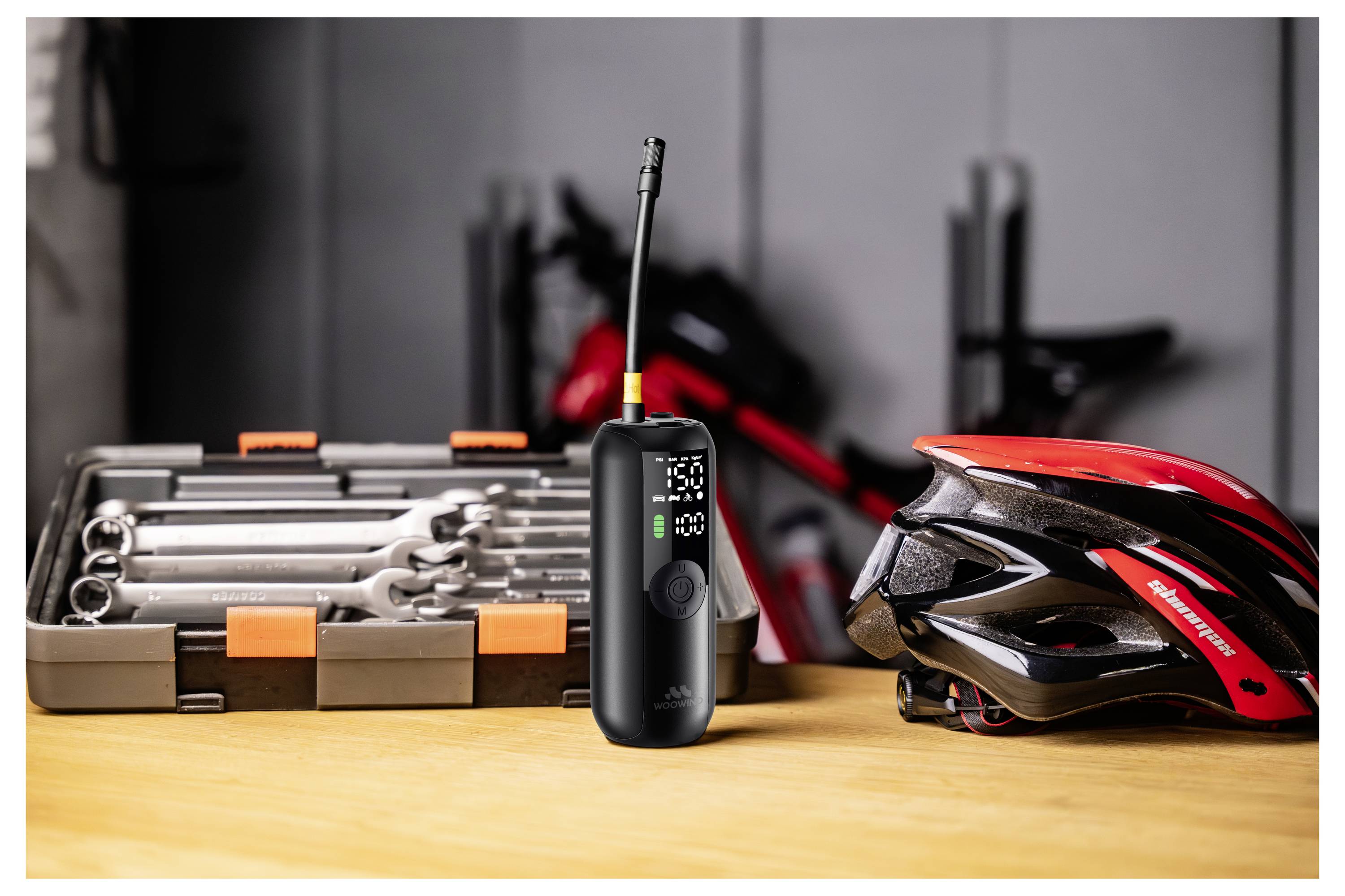 En trådlös luftpump på ett bord bredvid en röd cykelhjälm och en verktygslåda med skiftnycklar, vilket indikerar cykelunderhållsverktyg.