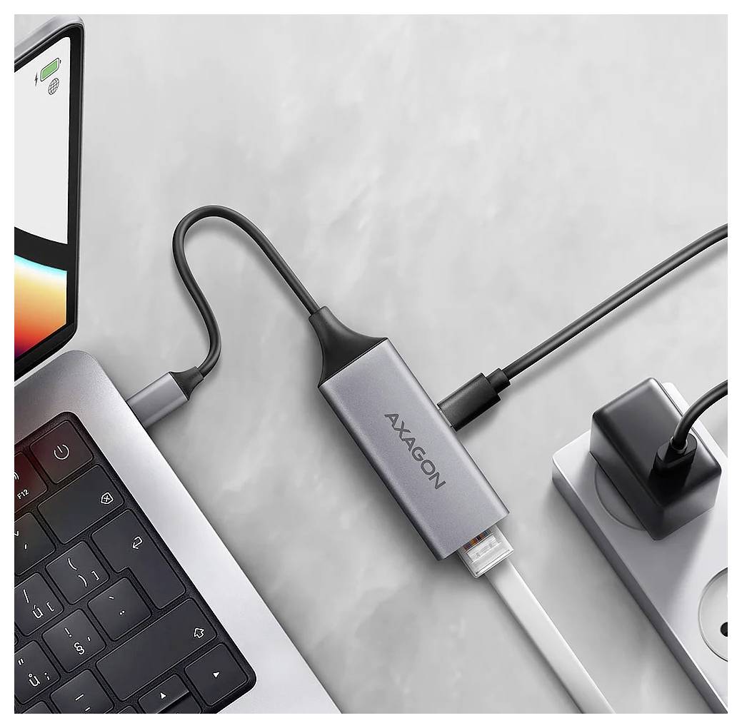 En bärbar dator ansluten till en AXAGON USB-C-hubb med flera kablar och en strömadapter på en ljusgrå yta.