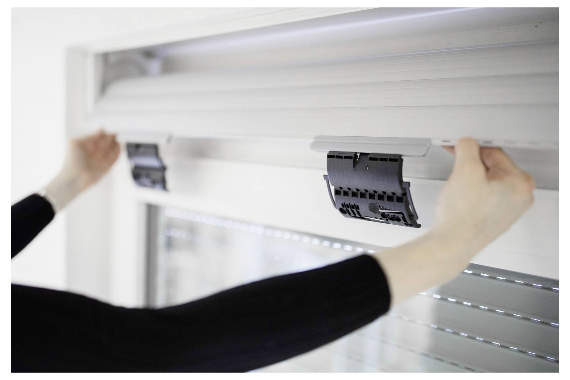 Person justerar en rullgardin högst upp på ett fönster, som visar mekanismen och positionen för installation eller underhåll.