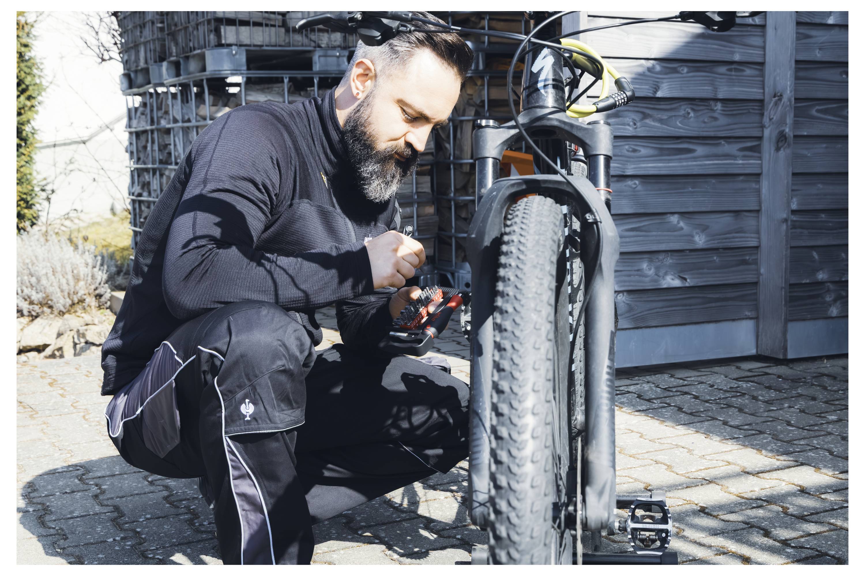 En man knäer bredvid en cykel och justerar växlarna med ett verktyg. Han är klädd i svart, fokuserad intensivt, utomhus på en solig dag.