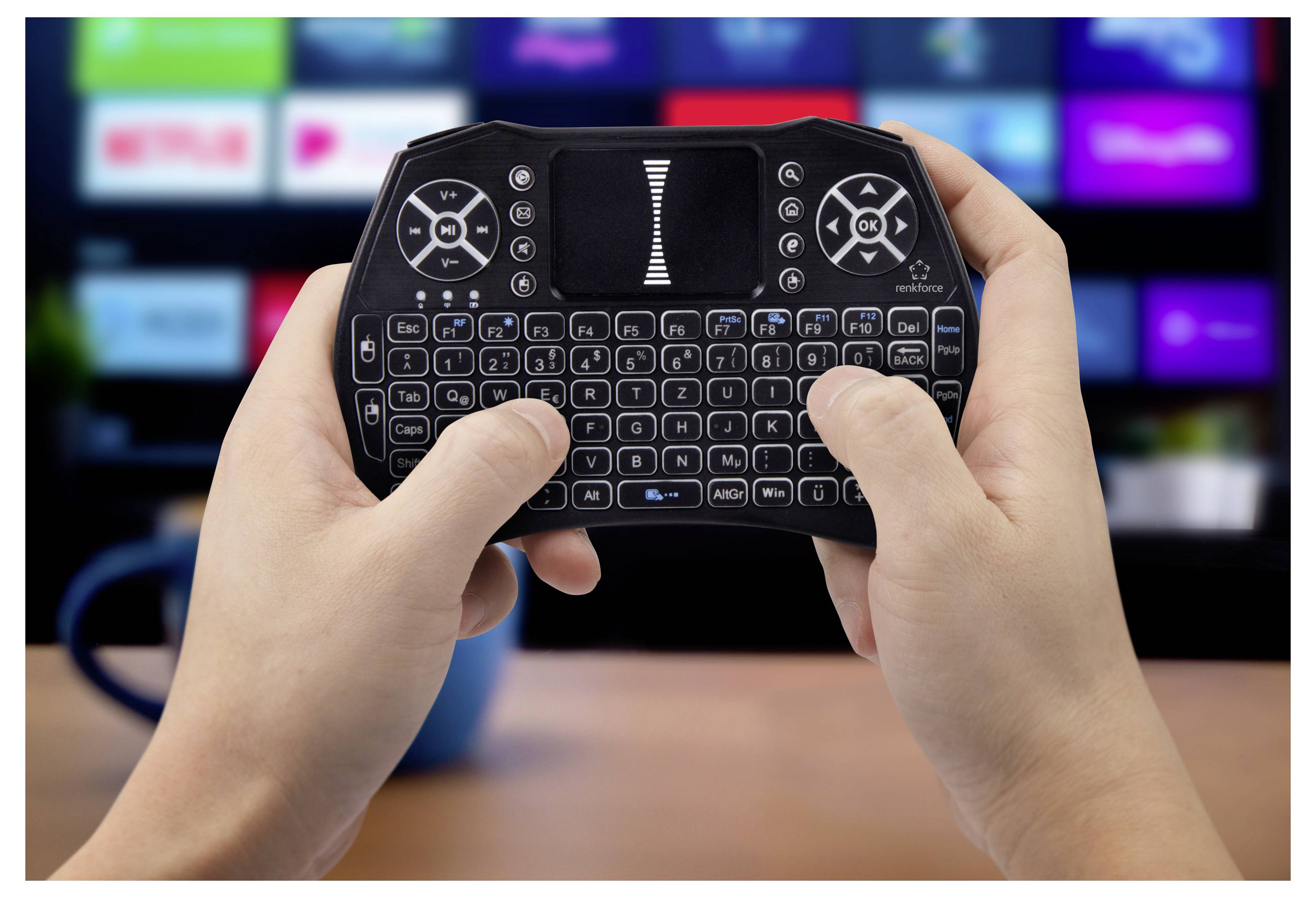 Händer som håller ett litet trådlöst tangentbord med pekplatta, framför en suddig TV-skärm, vilket antyder fjärrkontroll av media eller navigering av smart-TV.