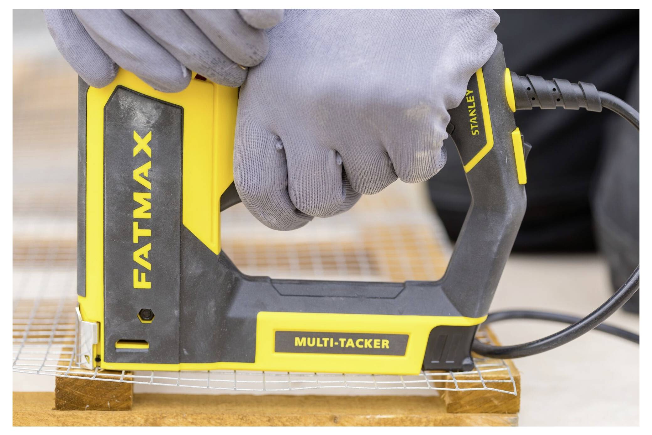 En person som bär handskar använder en gul och svart elektrisk häftapparat märkt "Fatmax Multi-Tacker" på ett nätmaterial över trä.