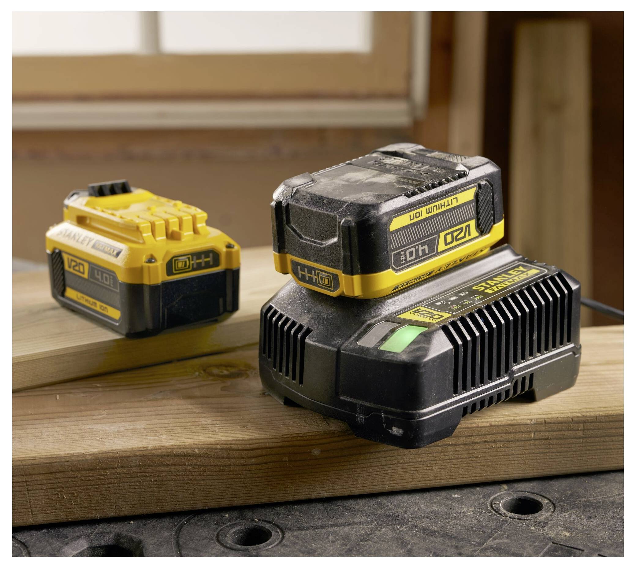 Två uppladdningsbara batterier, ett dockat på en laddare märkt "120V MAX", sitter på en träverkstad, vilket indikerar en elmaskinsuppsättning.