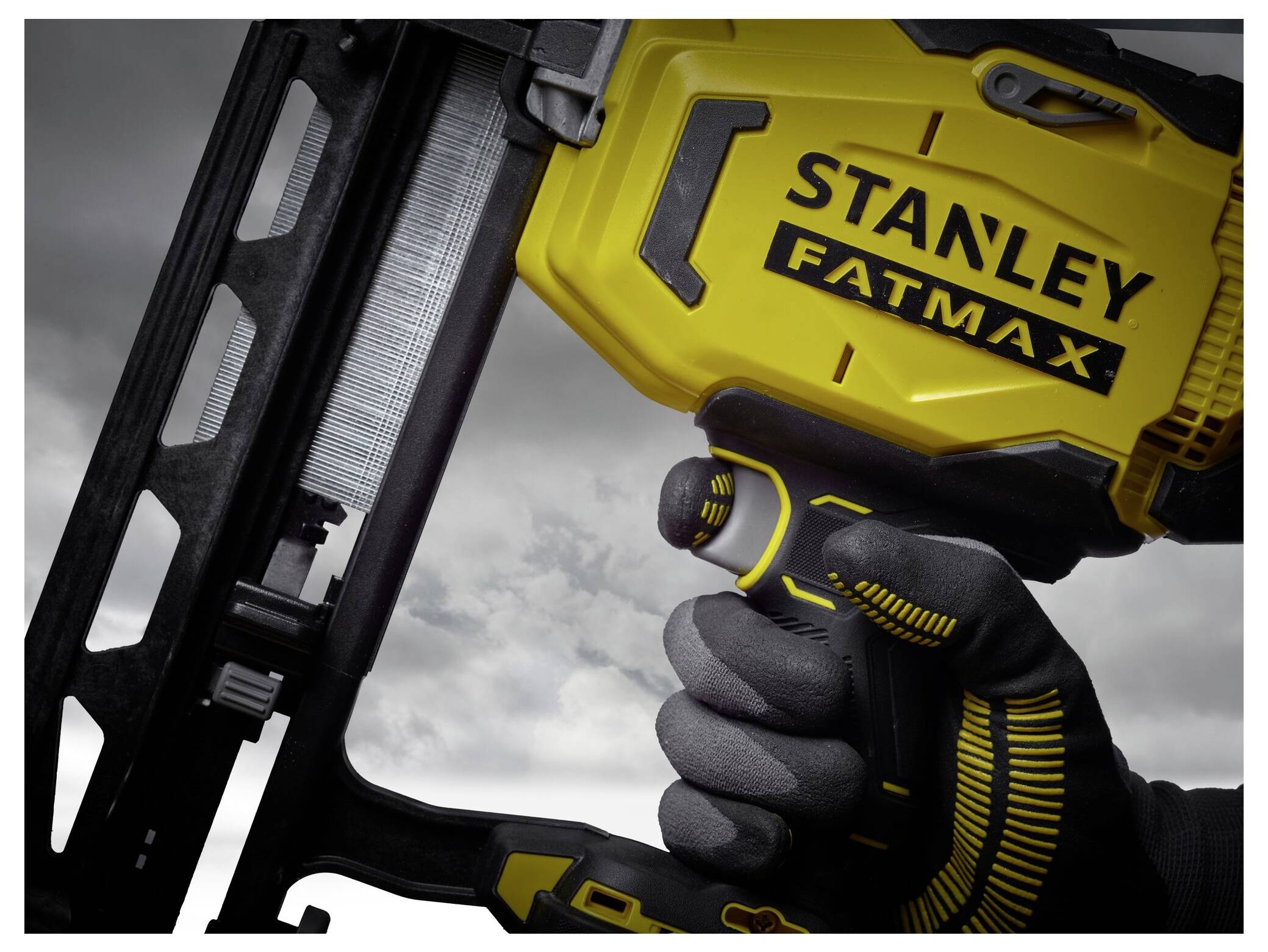 En person med handskar håller en gul Stanley Fatmax spikpistol mot en molnig himmel, vilket framhäver verktygets robusta design.