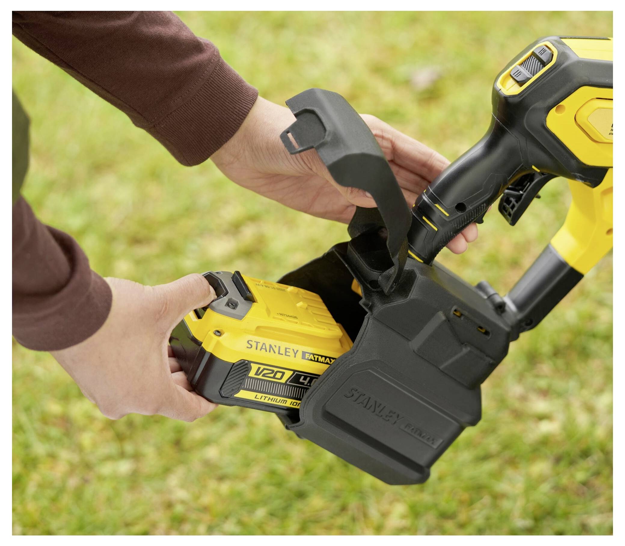 En person fäster ett gult "Stanley Fatmax V20"-batteri på ett elverktyg utomhus på en gräsig yta.