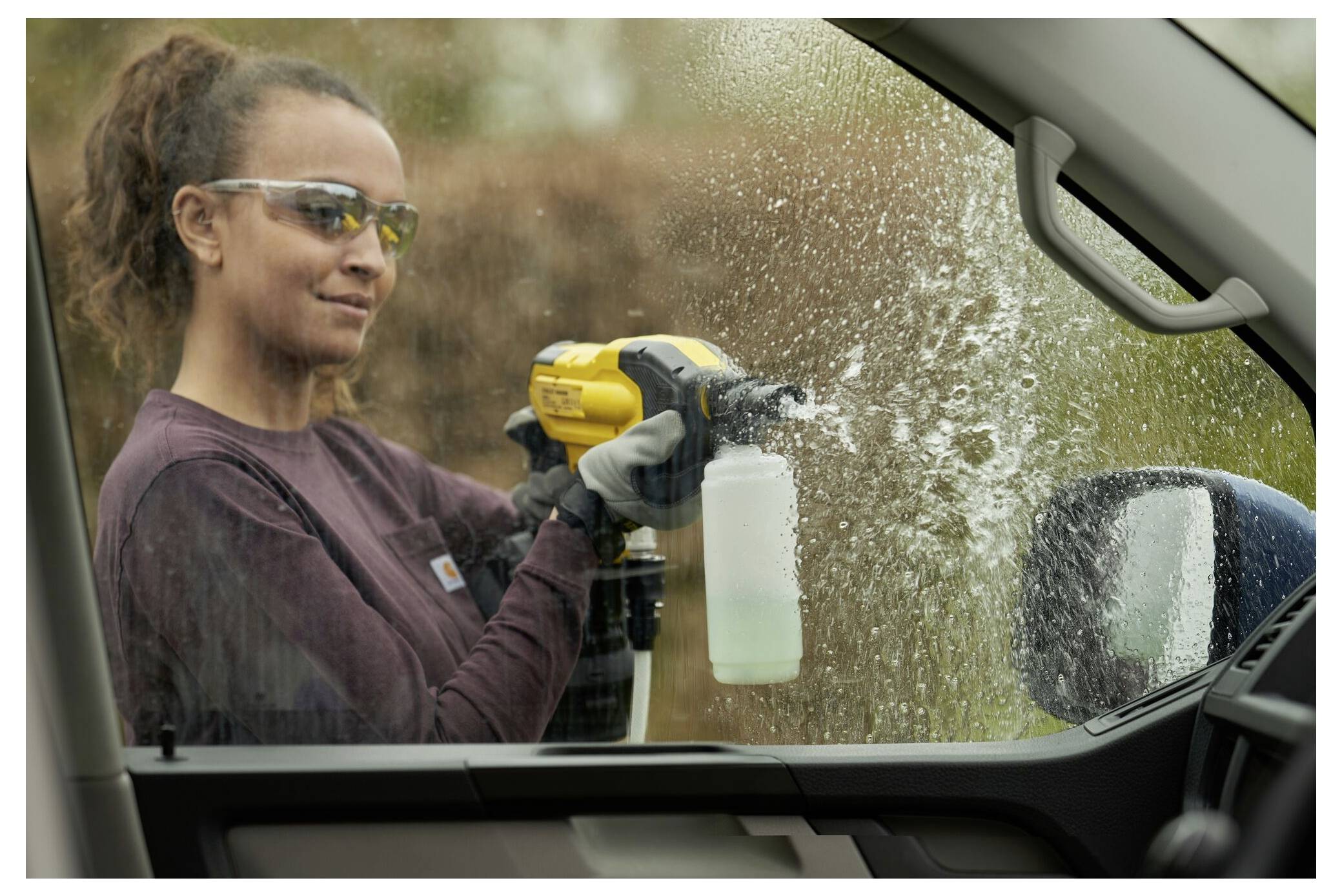 En person som bär skyddande glasögon och handskar använder ett motoriserat sprayverktyg för att rengöra ett bilfönsteri från utsidan.