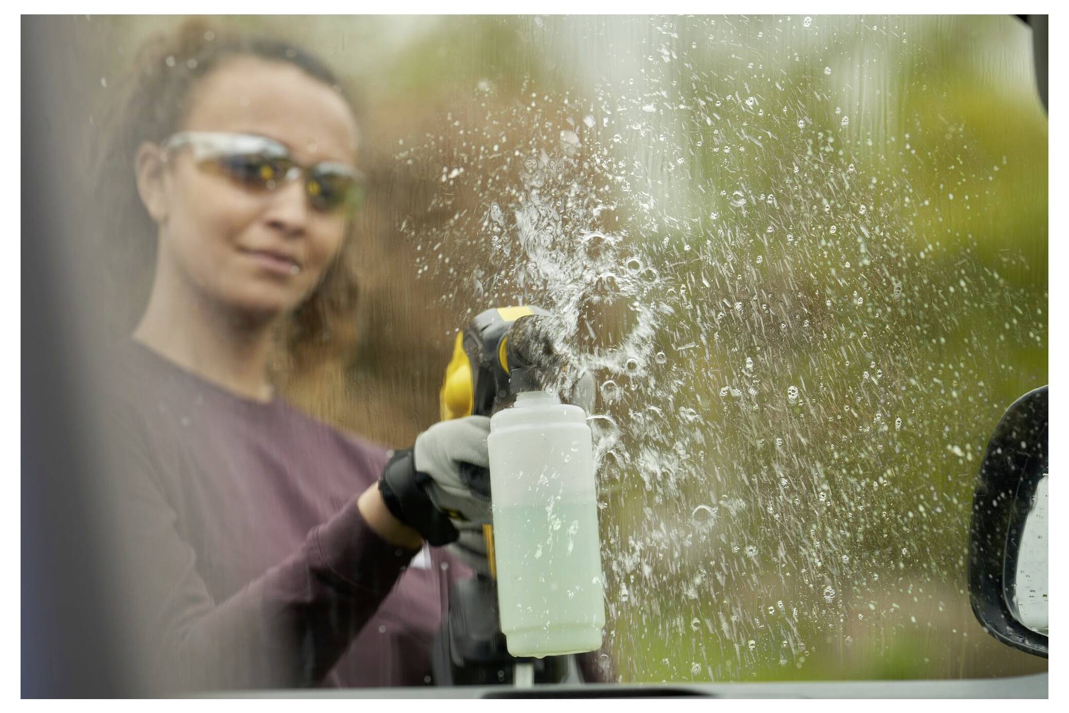 En person som bär skyddande solglasögon rensar ett fönster med en sprayflaska, vilket skapar synliga droppar på glasYtan.