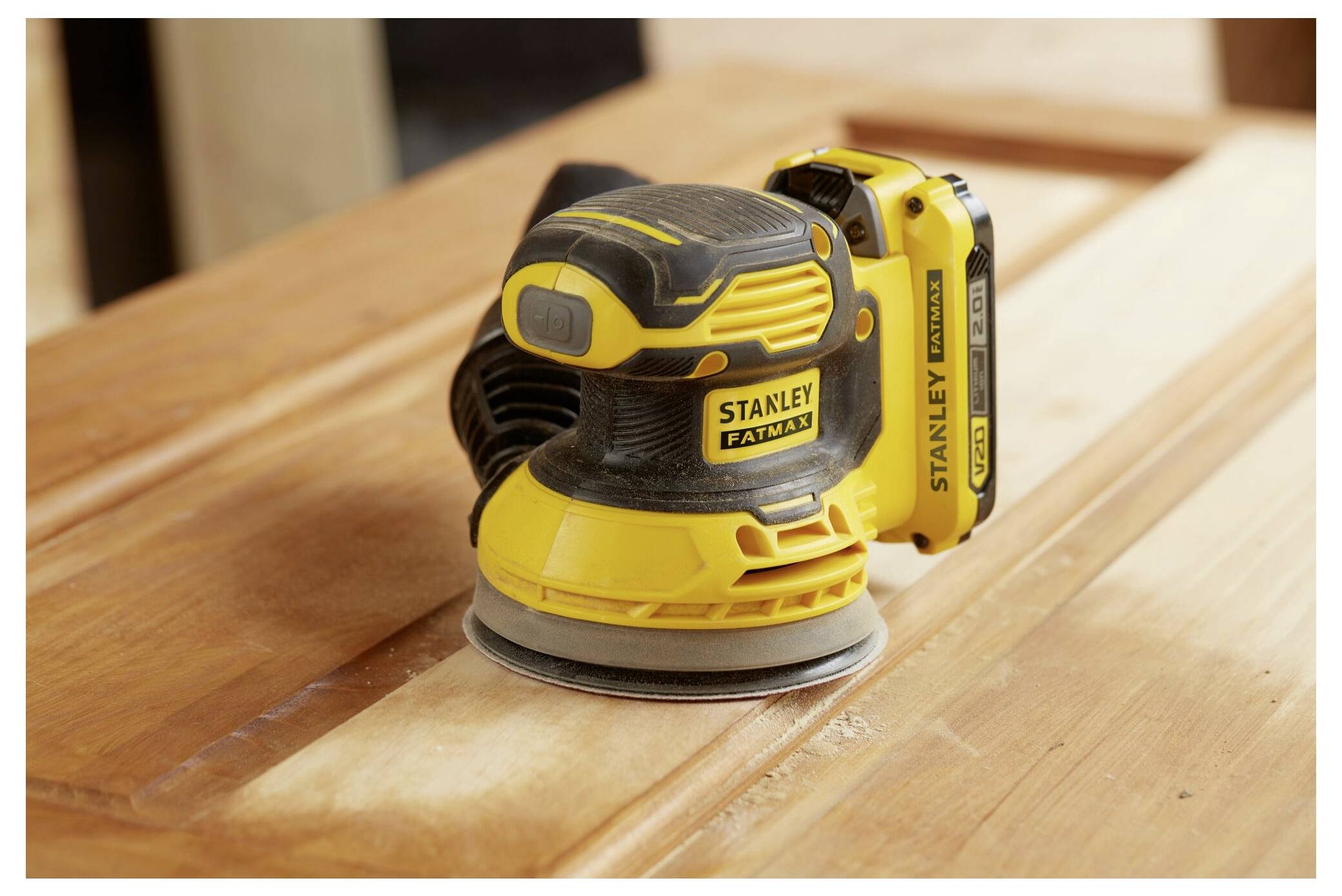 En gul och svart Stanley FatMax trådlös excenterslipar står på en träyta och framhäver dess ergonomiska design och batteriplaceringen.