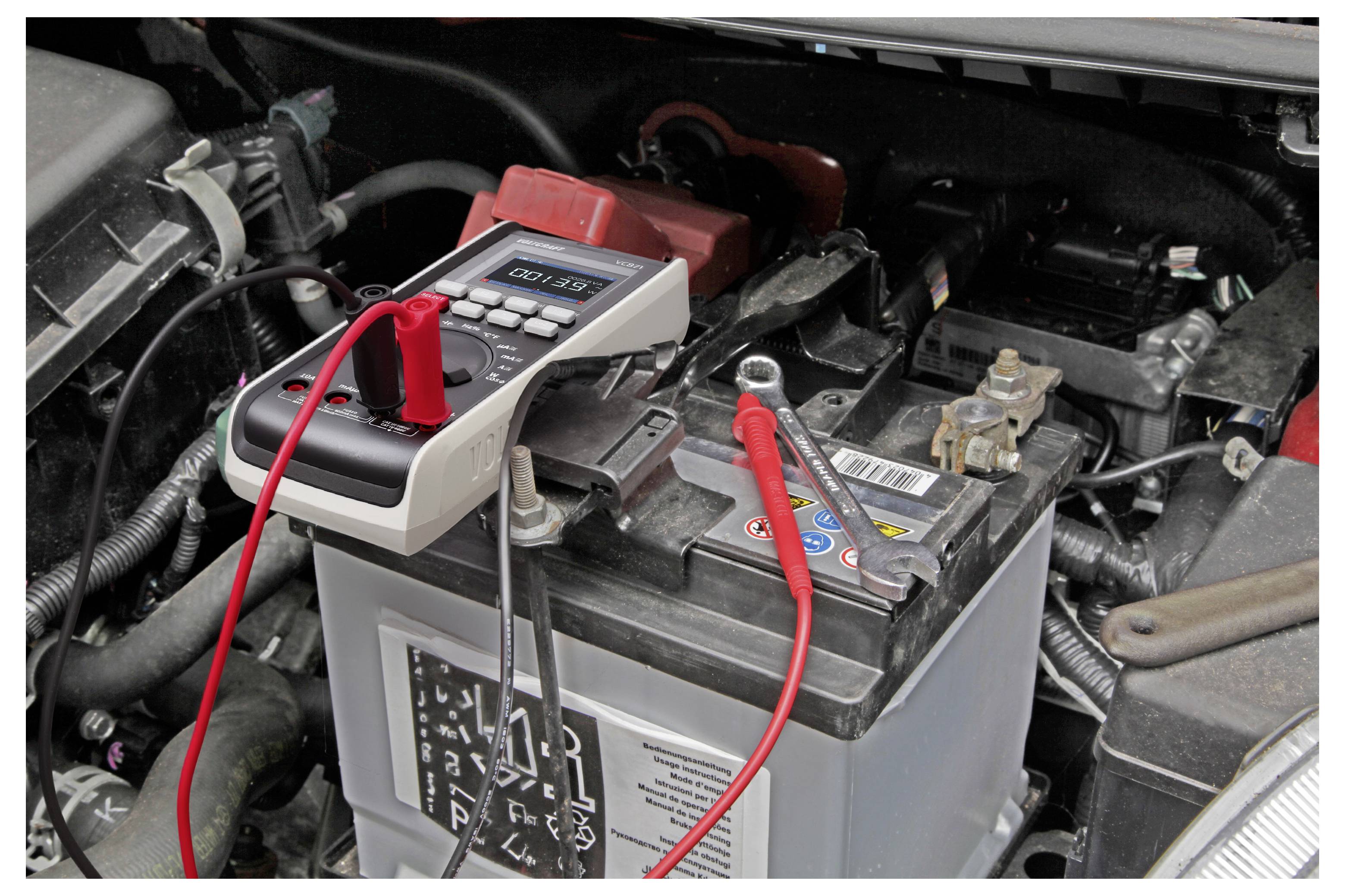 Multimeter som testar ett bilbatteri i motorutrymmet med röda och svarta ledningar anslutna till batteripolerna.