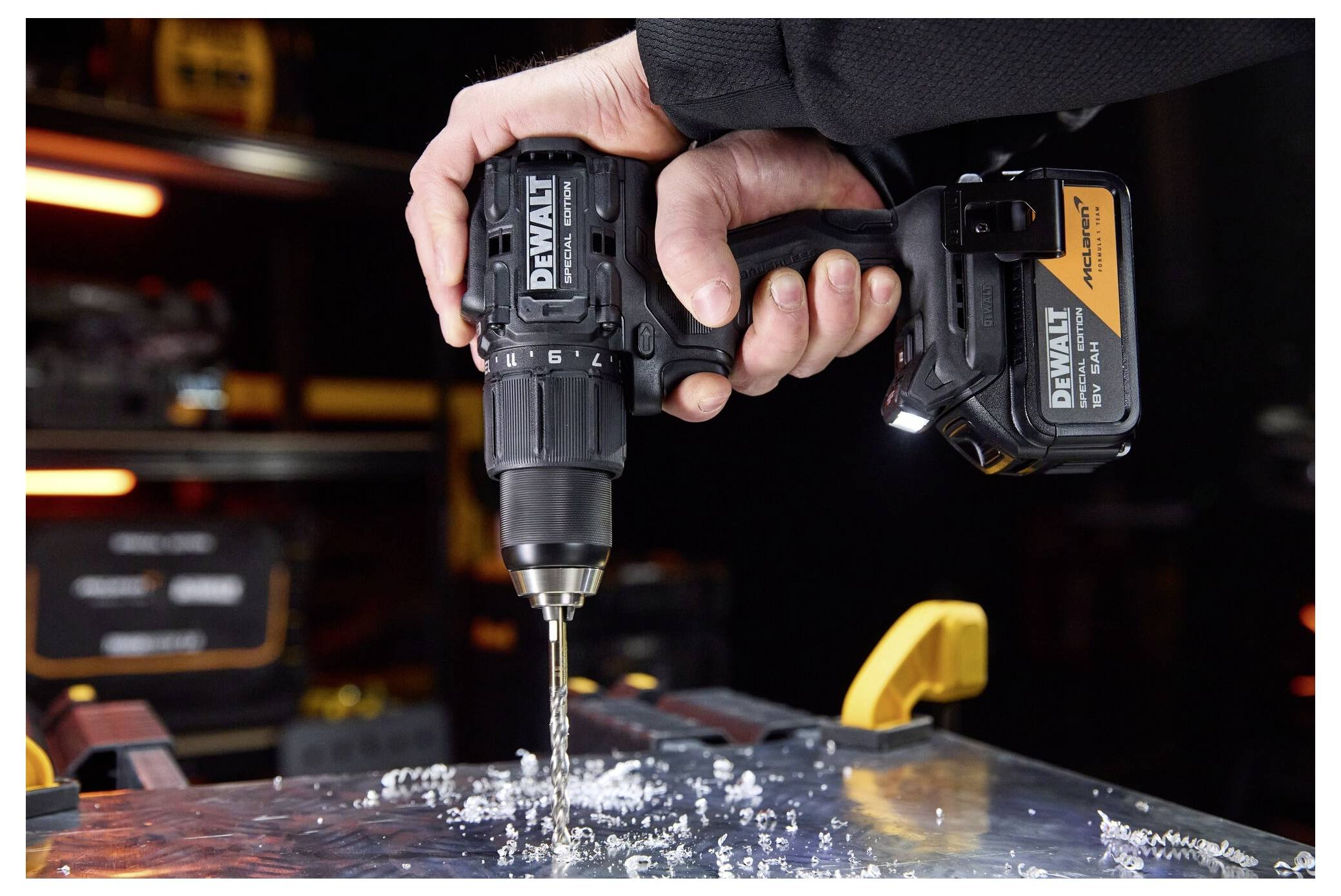 Hand som håller en DEWALT trådlös borrmaskin med borr som borrar in i metall och skapar spån; verktygsbatteri synligt i en verkstadsmiljö.