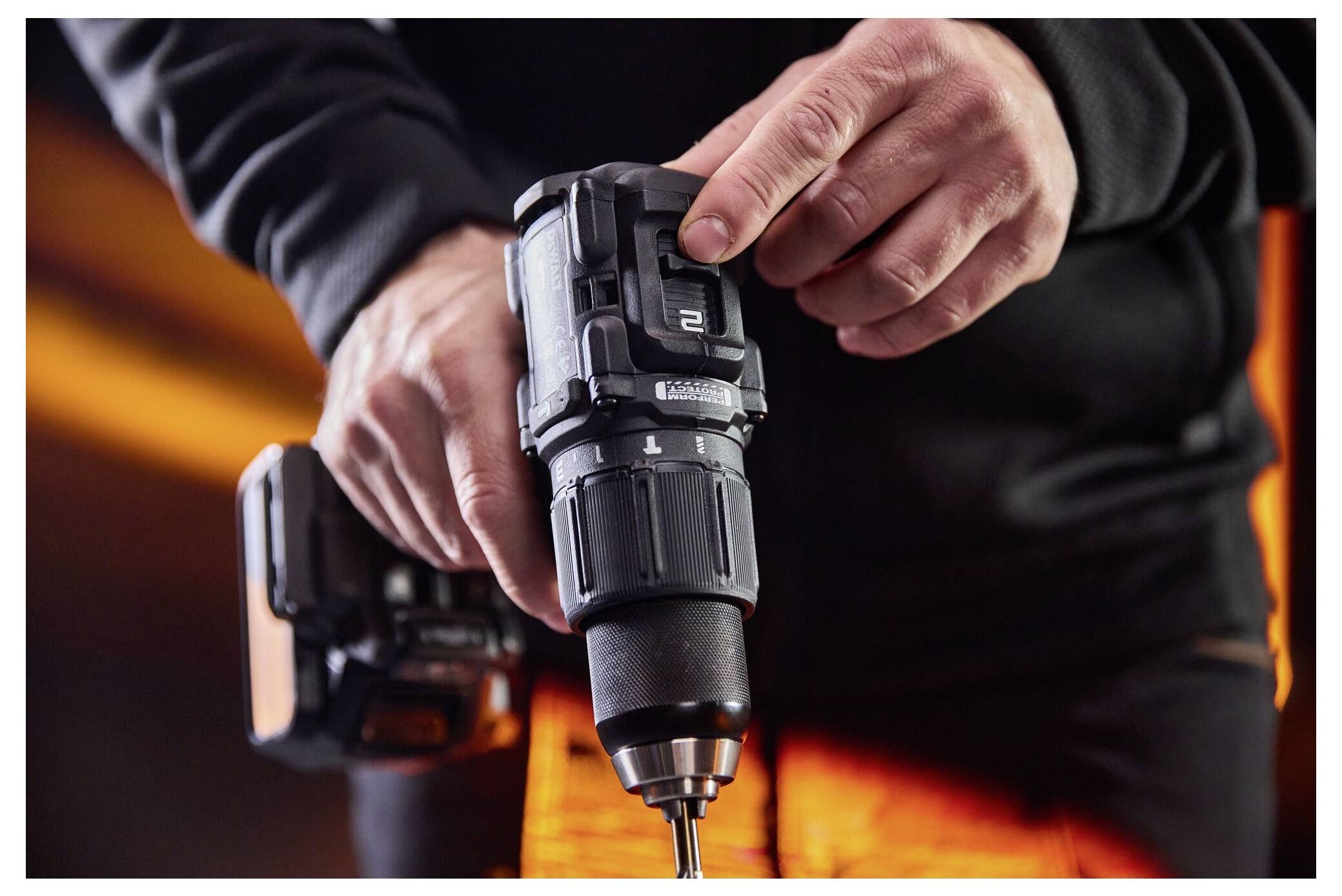 En person använder en trådlös borrmaskin, håller och strammar en borrbits, med ett batteribatteri anslutet och orange arbetskläder i bakgrunden.