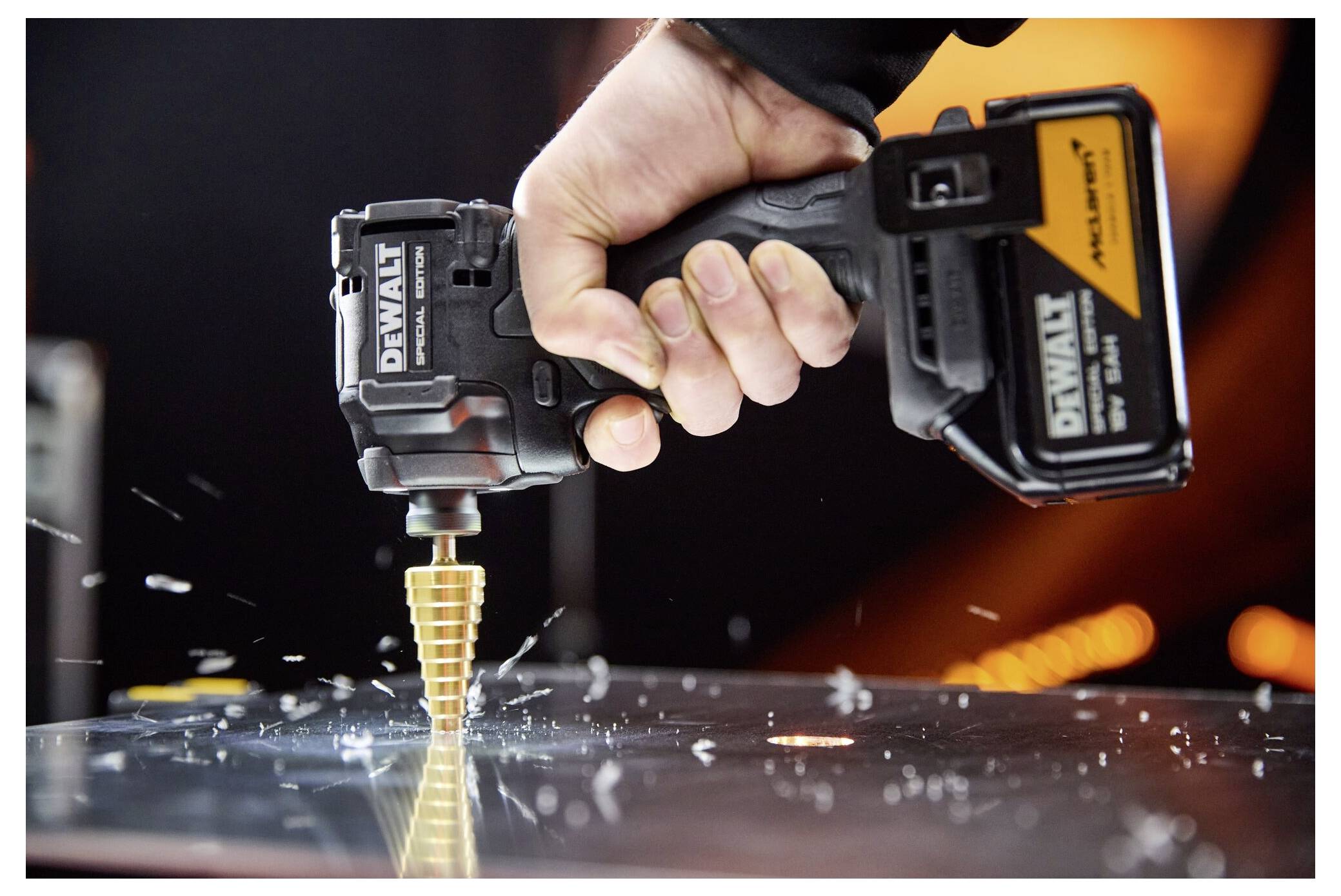 En hand håller en DeWalt trådlös borr som driver en guldpläterad stegvis borr in i ett metallark, med gnistor och metallspån som flyger omkring.