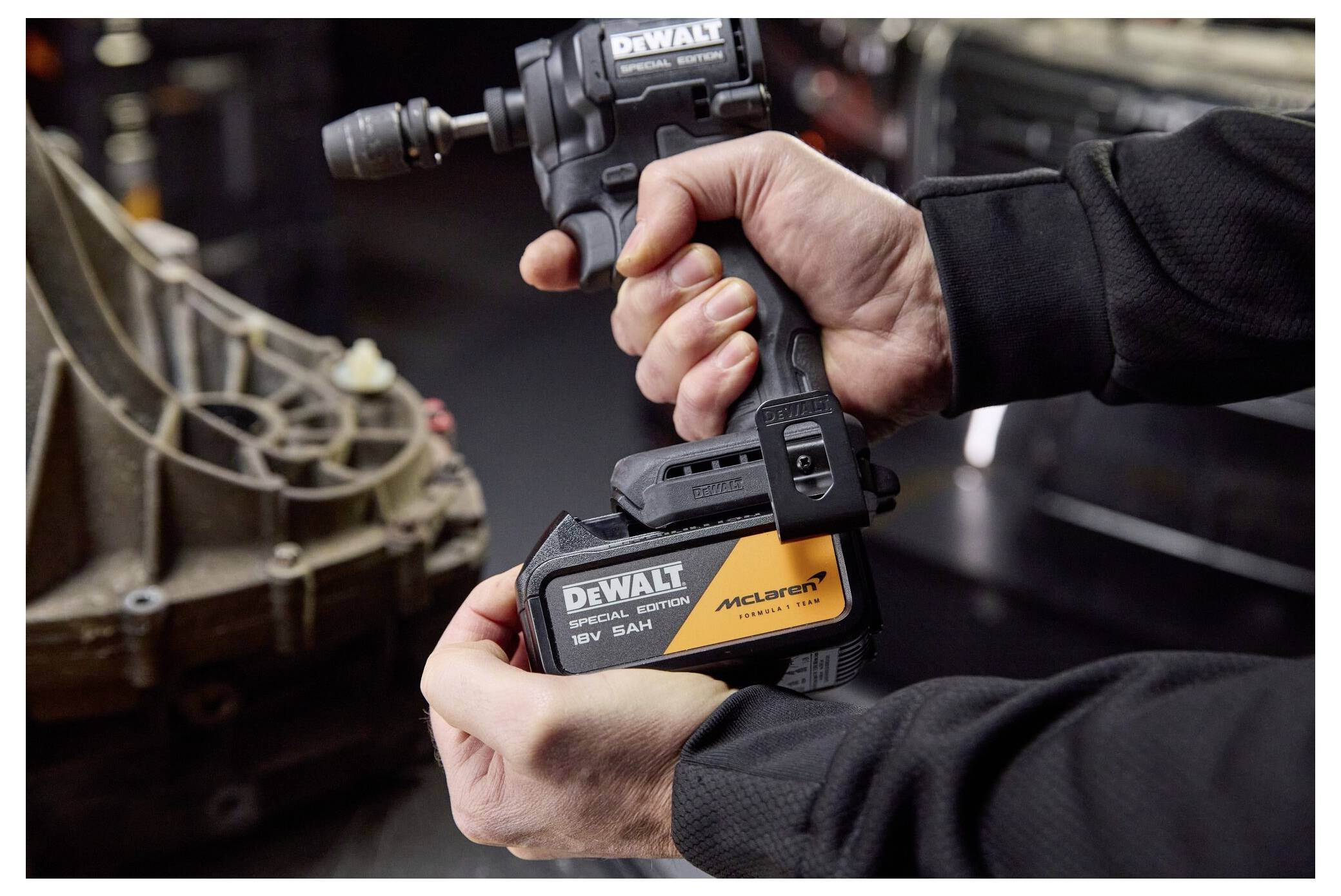 Närbild av händer som använder en DeWalt 18V trådlös slagskruvdragare med McLaren-batteri för att dra åt eller lossa en fordonsdetalj i ett garage.