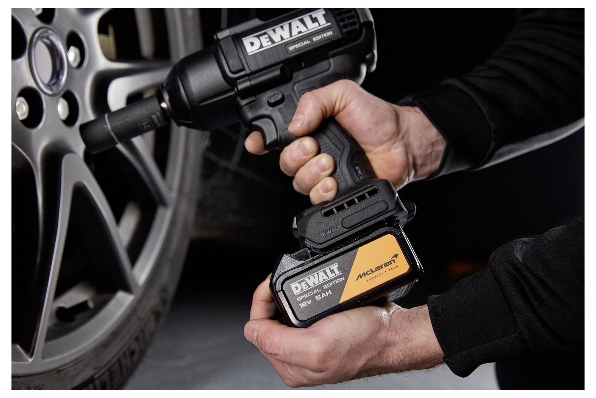 Händer som håller en DEWALT trådlös slagskruvdragare och batterisats bredvid ett bilhjul, som visar verktygs- och batteriinstallation.