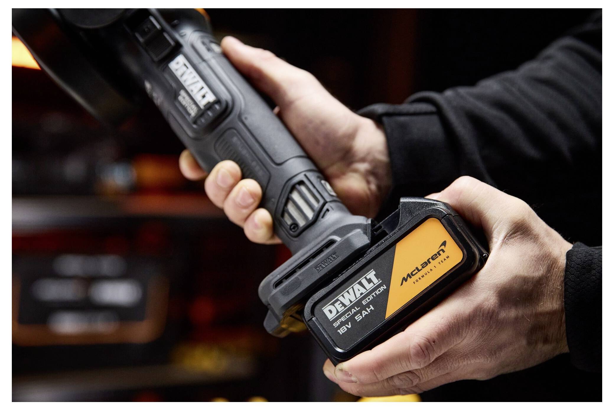 Händer som håller ett DeWalt trådlöst verktygsackupaket märkt "McLaren" och "DEWALT", visad i närbild i en verkstadsmiljö.