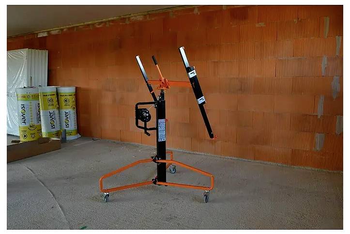 Orange-och-svart gipsplåtshiss stående på hjul i ett oavslutad rum med exponerade tegelväggar och byggmaterial.