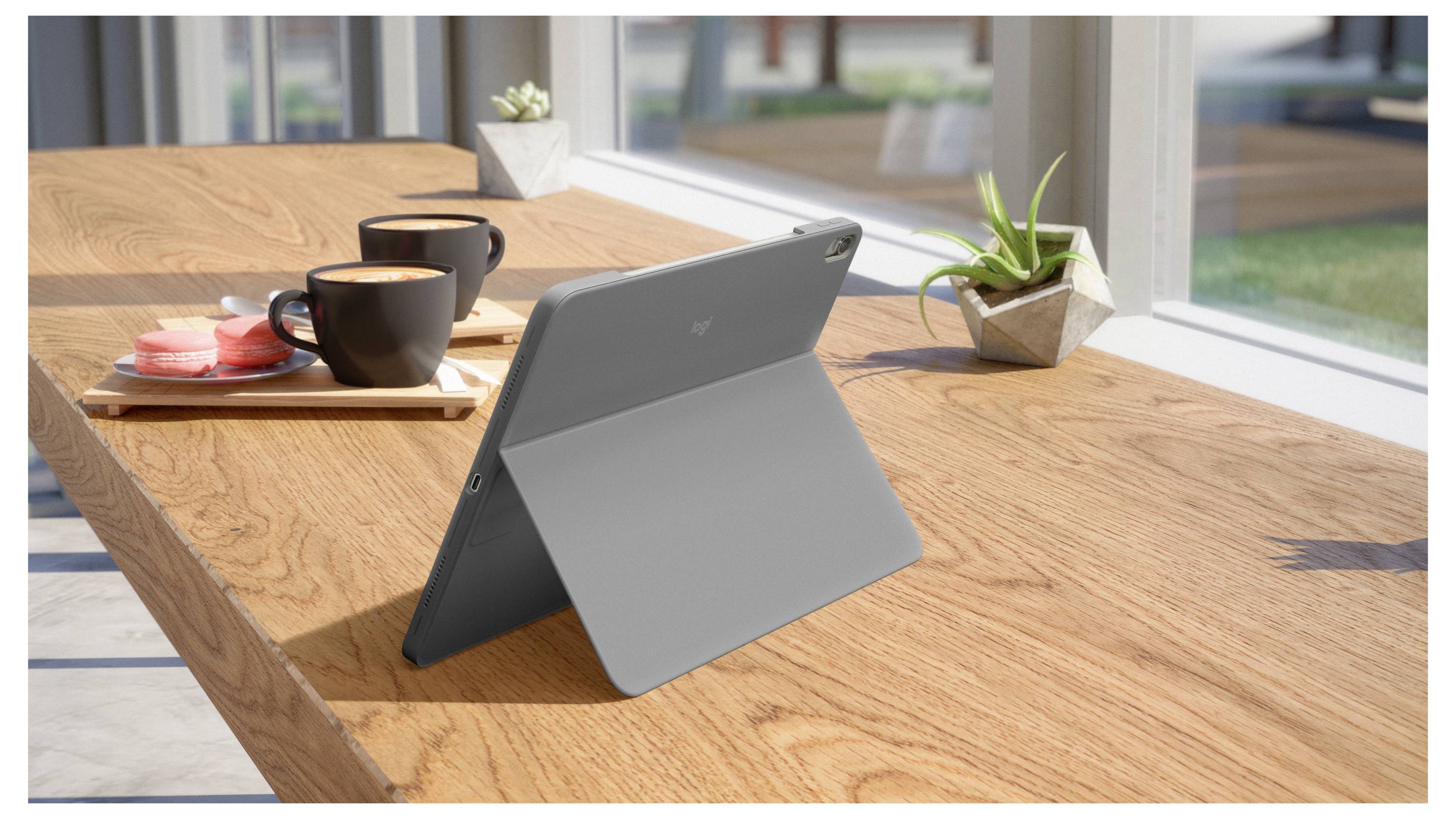 Tablet i grå foliohållare på ett träbord vid ett fönster, bredvid två kaffekoppar, en bricka med bakverk och små krukväxter.