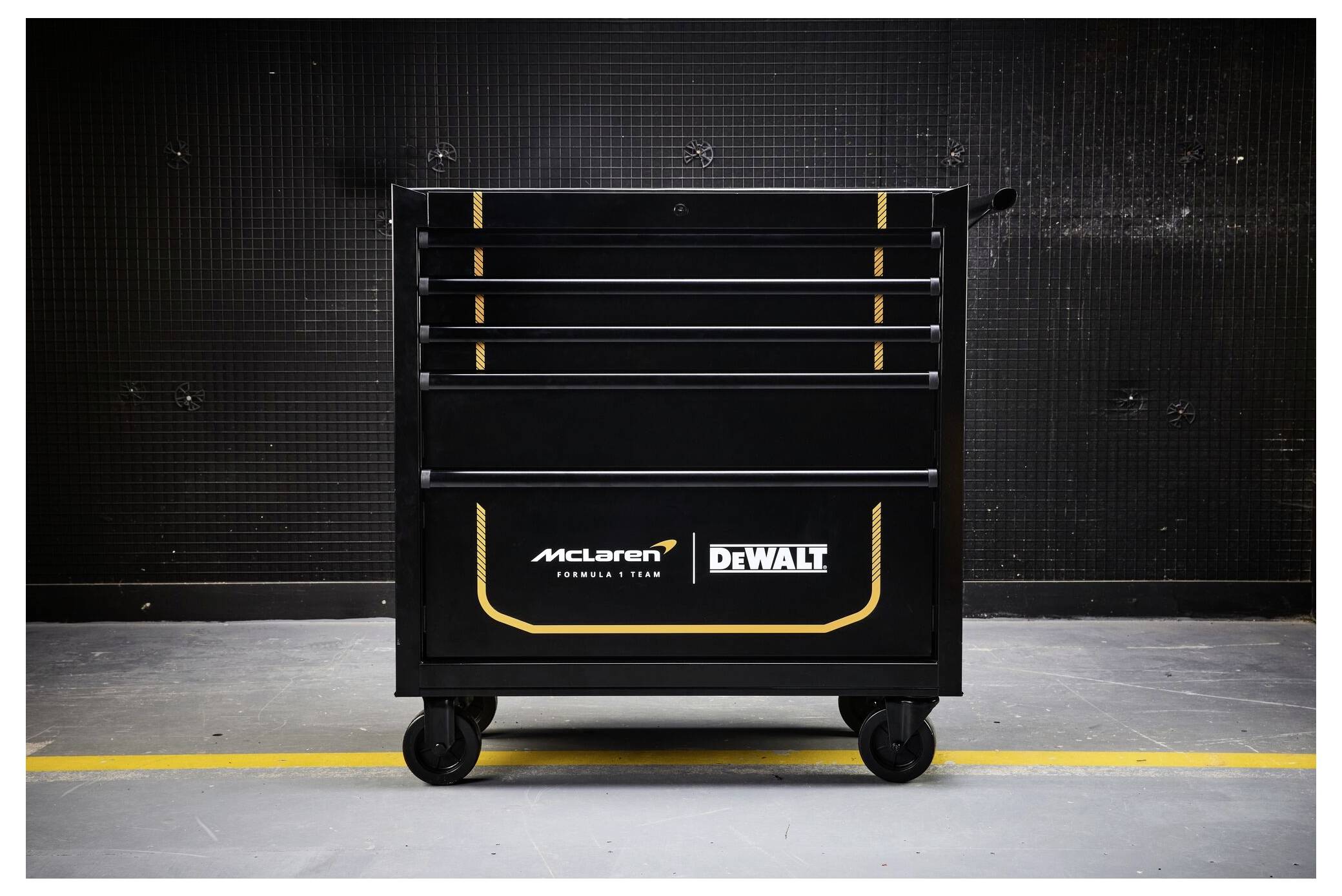 Svart verktygsväska med hjul, med McLaren- och DEWALT-logotyper, centrerad mot en mörk industrivägg.