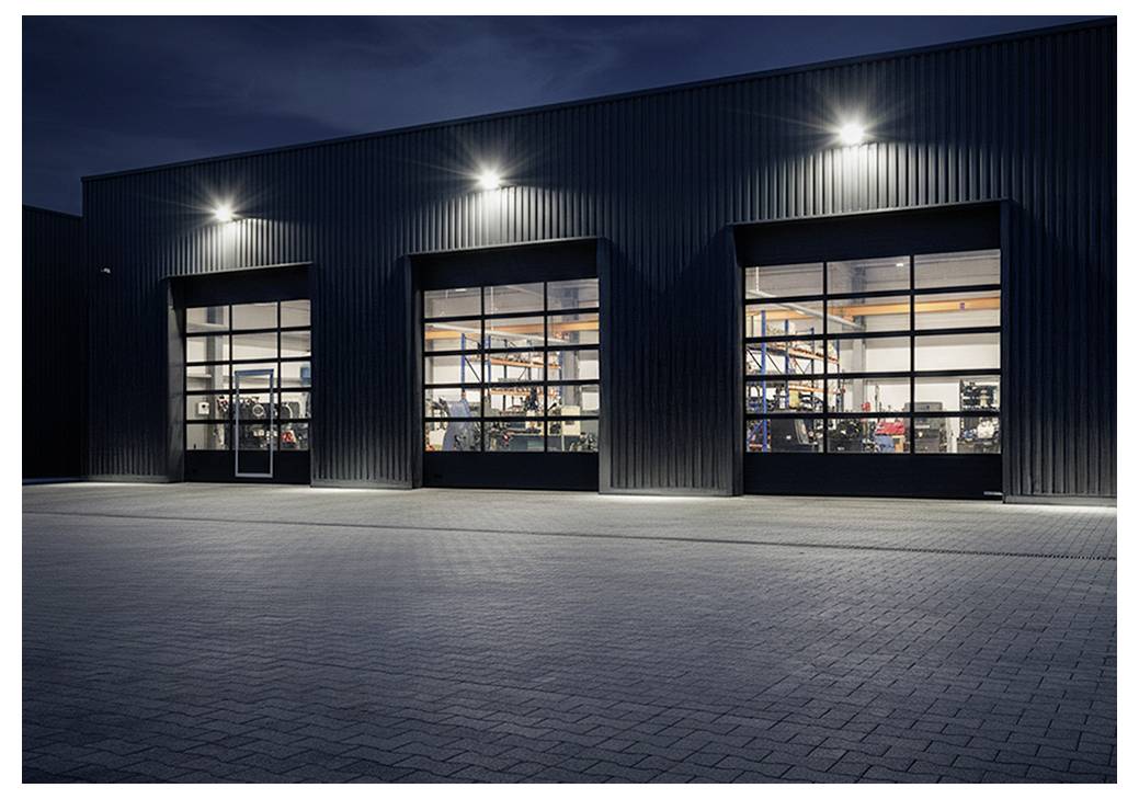 Exteriör av ett modernt industriellt lager på natten, med tre stora upplysta garagedörrar som visar utrustning inomhus.