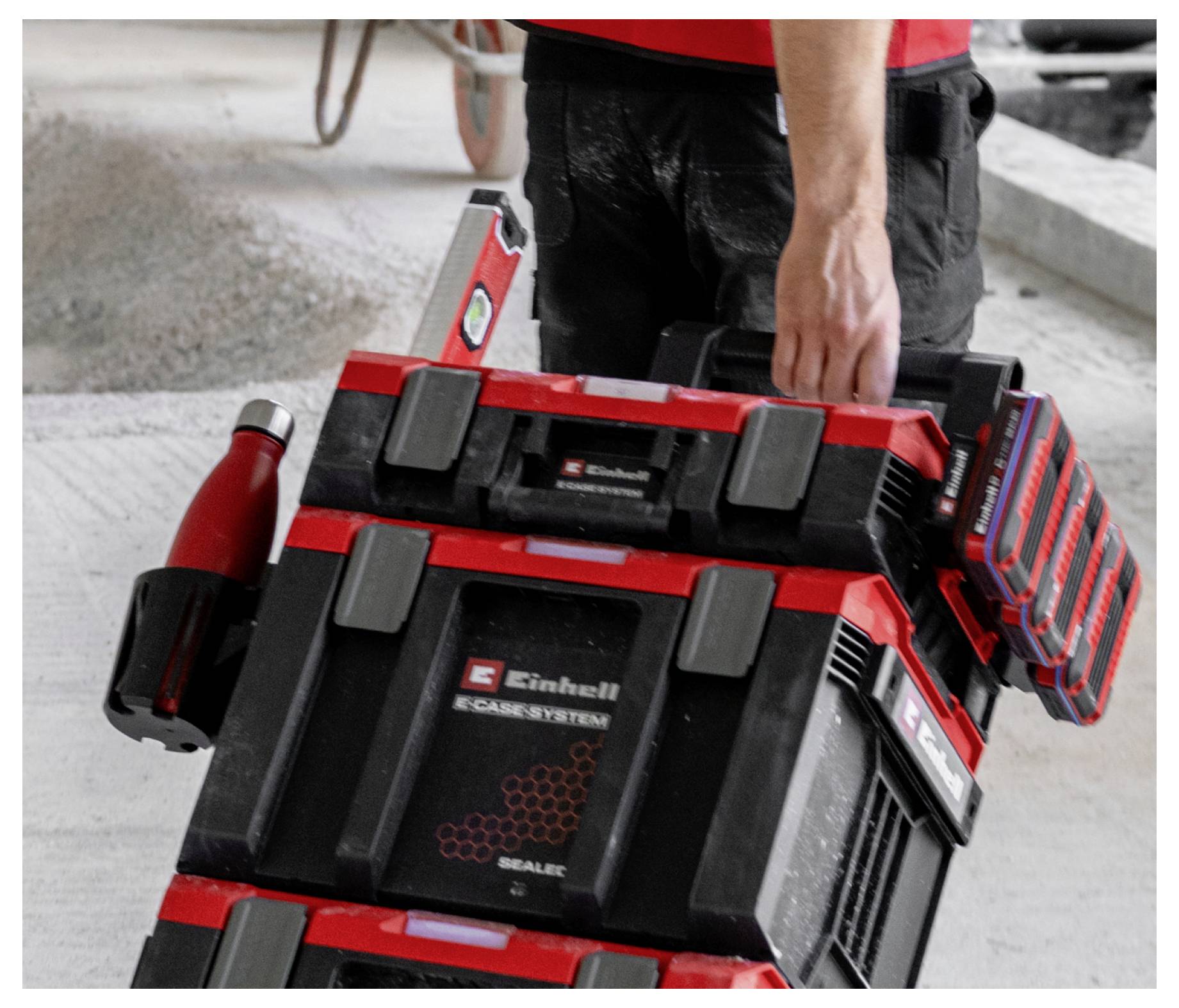 Arbetare som bär staplade röd-och-svarta Einhell-verktygslådor och ett trådlöst batteriladdningspaket på en byggnadsplats.