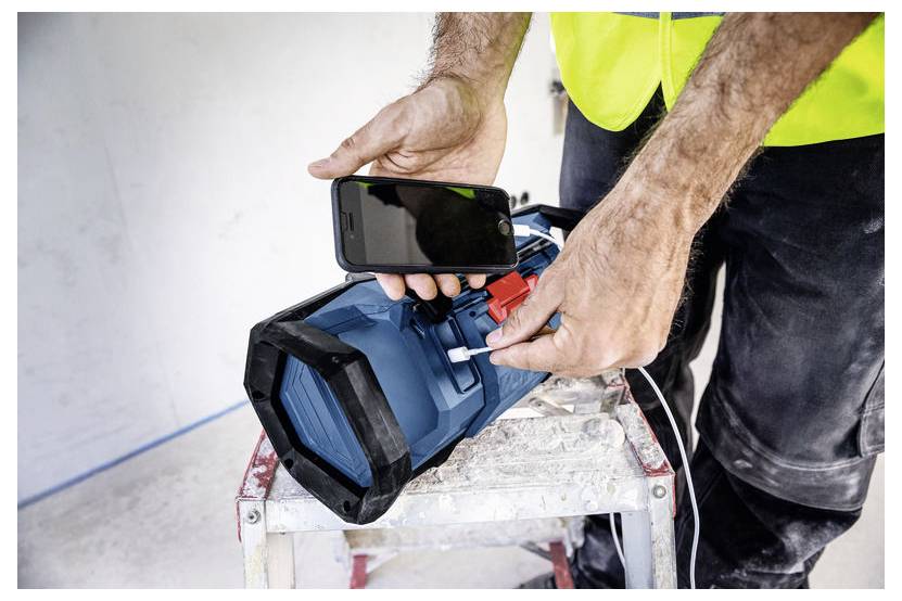 En person i en highvis-väst använder en smartphone för att ladda eller ansluta en blå bärbar högtalare på en arbetsbänk.