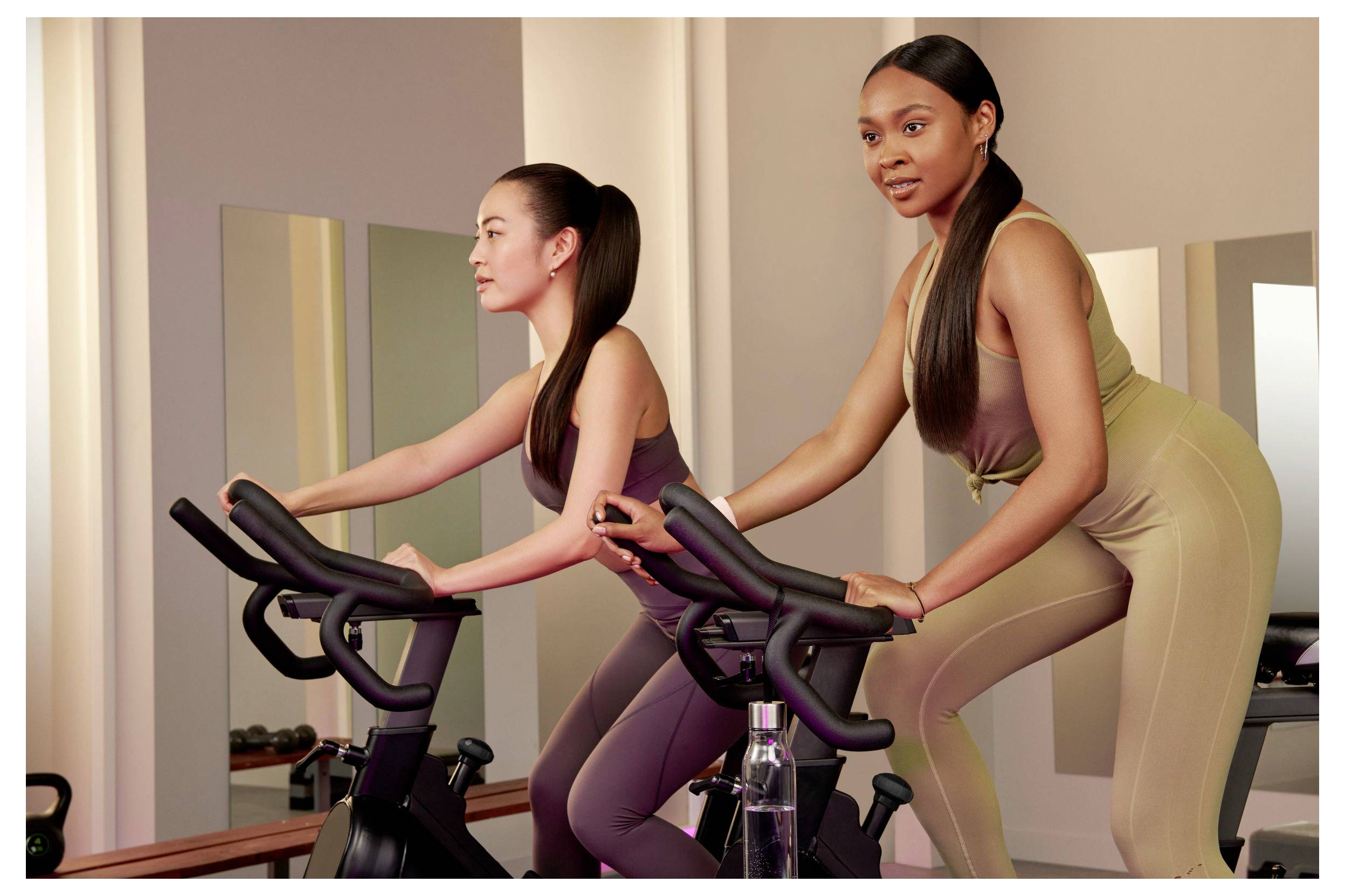 Två kvinnor som tränar på motionscyklar i en gymstudio, en framför och en bakom, båda lutar sig framåt och trampar.
