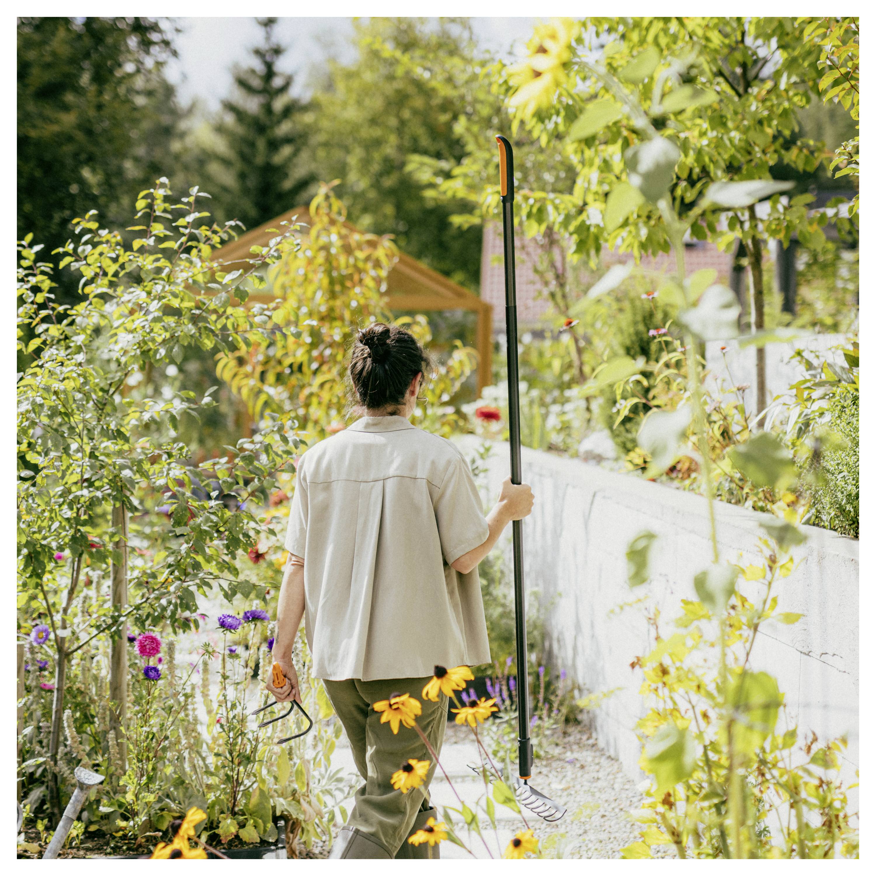 En person arbetar i trädgården i en frodig bakgård, holding ett långskaftat verktyg bredvid en blomsterfylld väg och en vit vägg nära ett litet skjul.