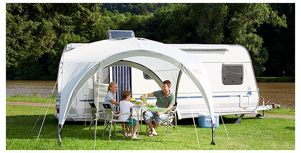 En familj som sitter vid ett bord under ett överdrag bredvid en vit husvagn på en campingplats, med träd och gräs i bakgrunden.