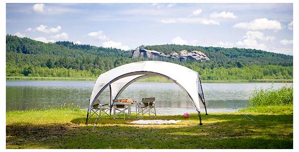 Vitt baldakintält med campingstolar och bord på en gräsbevuxen strandkant vid en sjö, med utsikt över lugnt vatten och skogsbeklädda kullar under en ljus himmel.