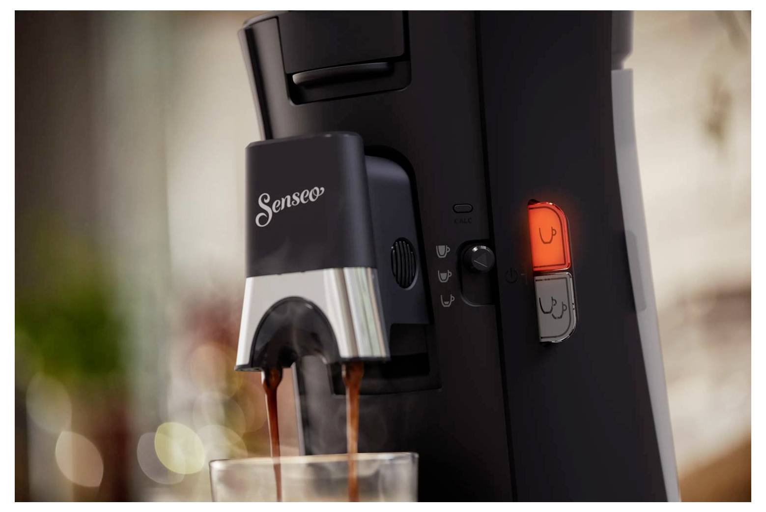 Närbild av en Senseo-kaffebryggare som dispenserar kaffe i ett glas, med den upplysta bryggknappen synlig på sidan.