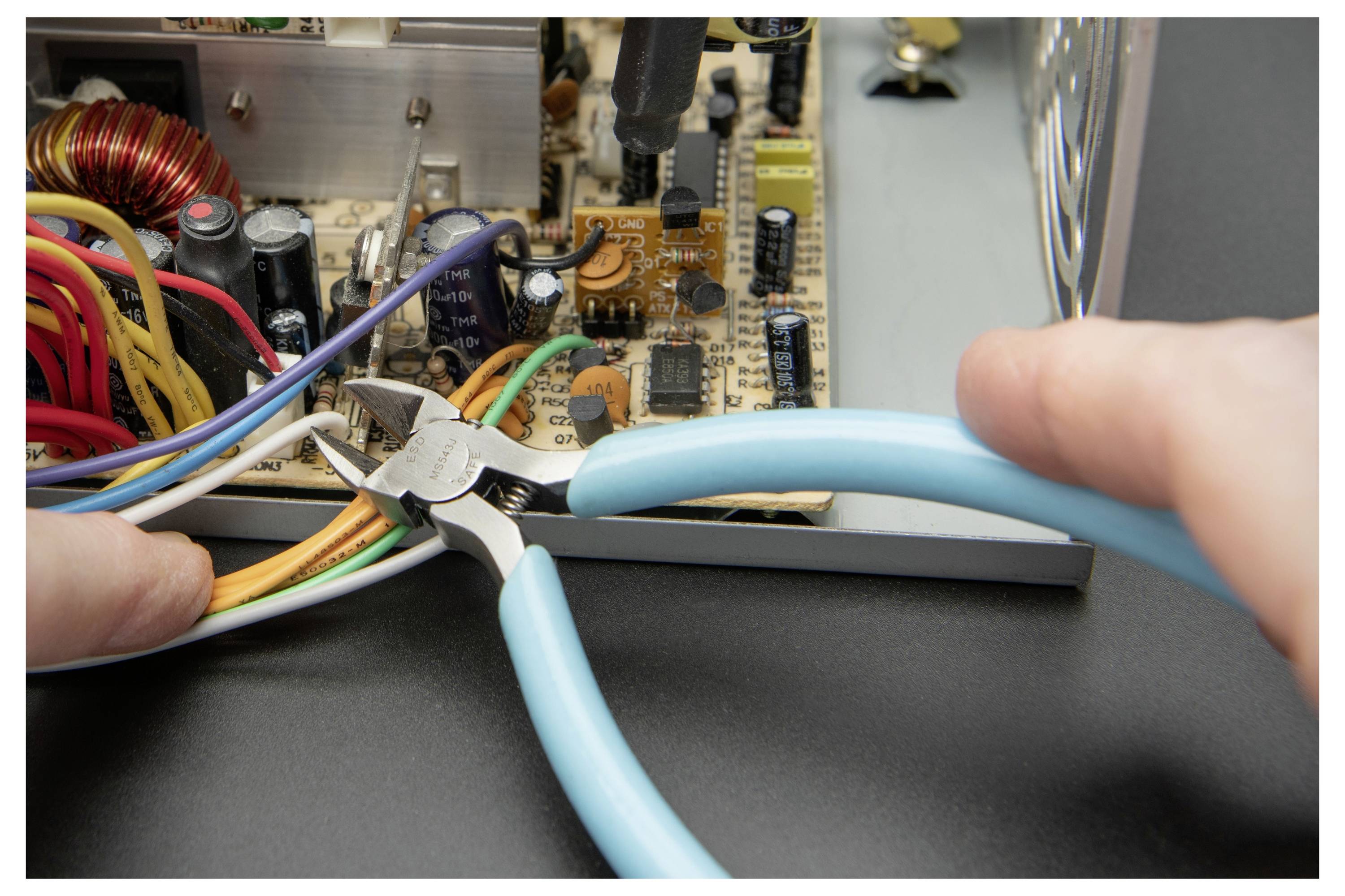 Händer använder blå-handtag tänger för att klippa ledningar inne i en öppen elektronisk strömförsyningsenhet.