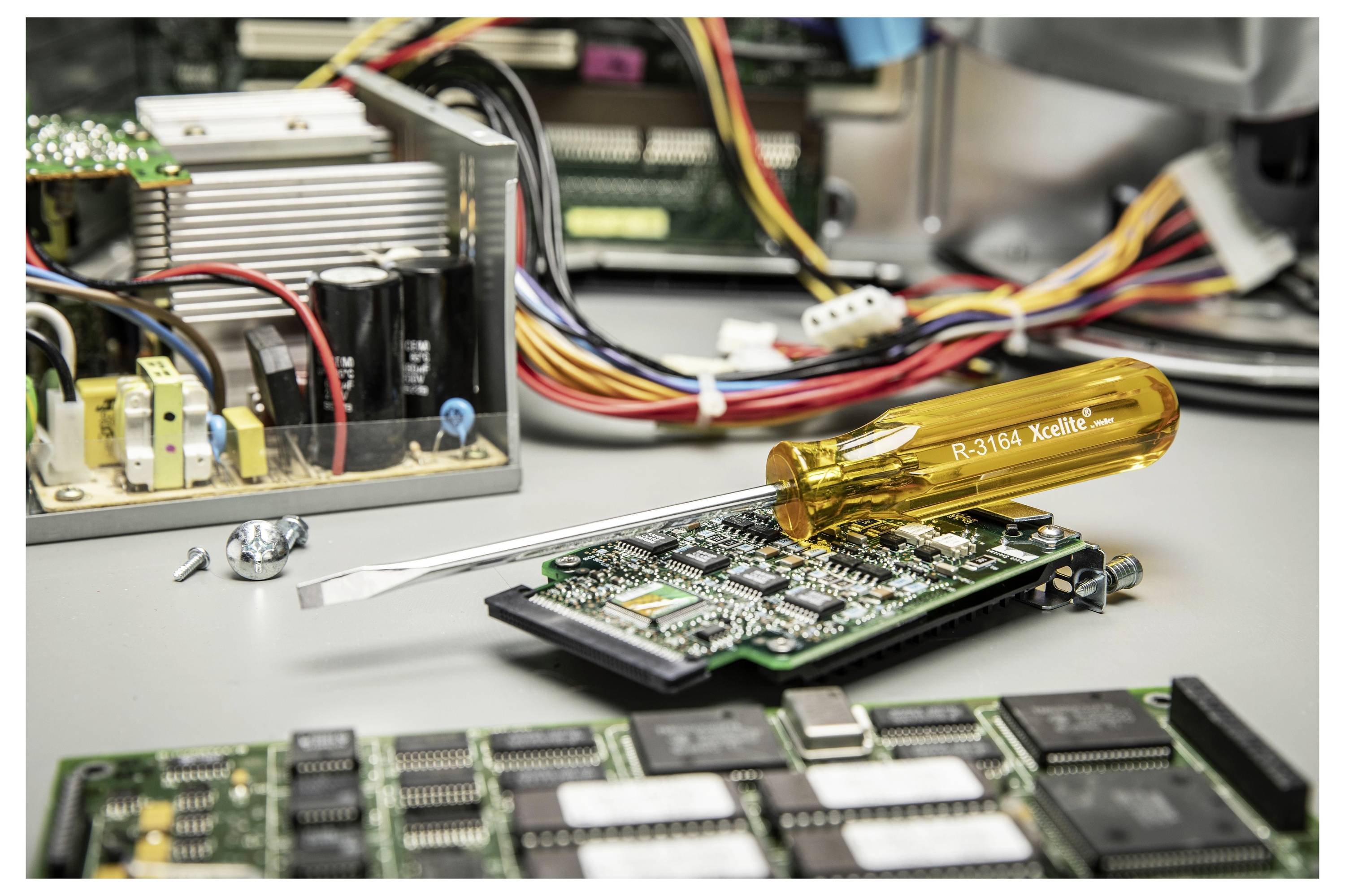 Elektronikreparationsbänk med en skruvmejsel på ett kretskort bredvid demonturade komponenter och datordelar.