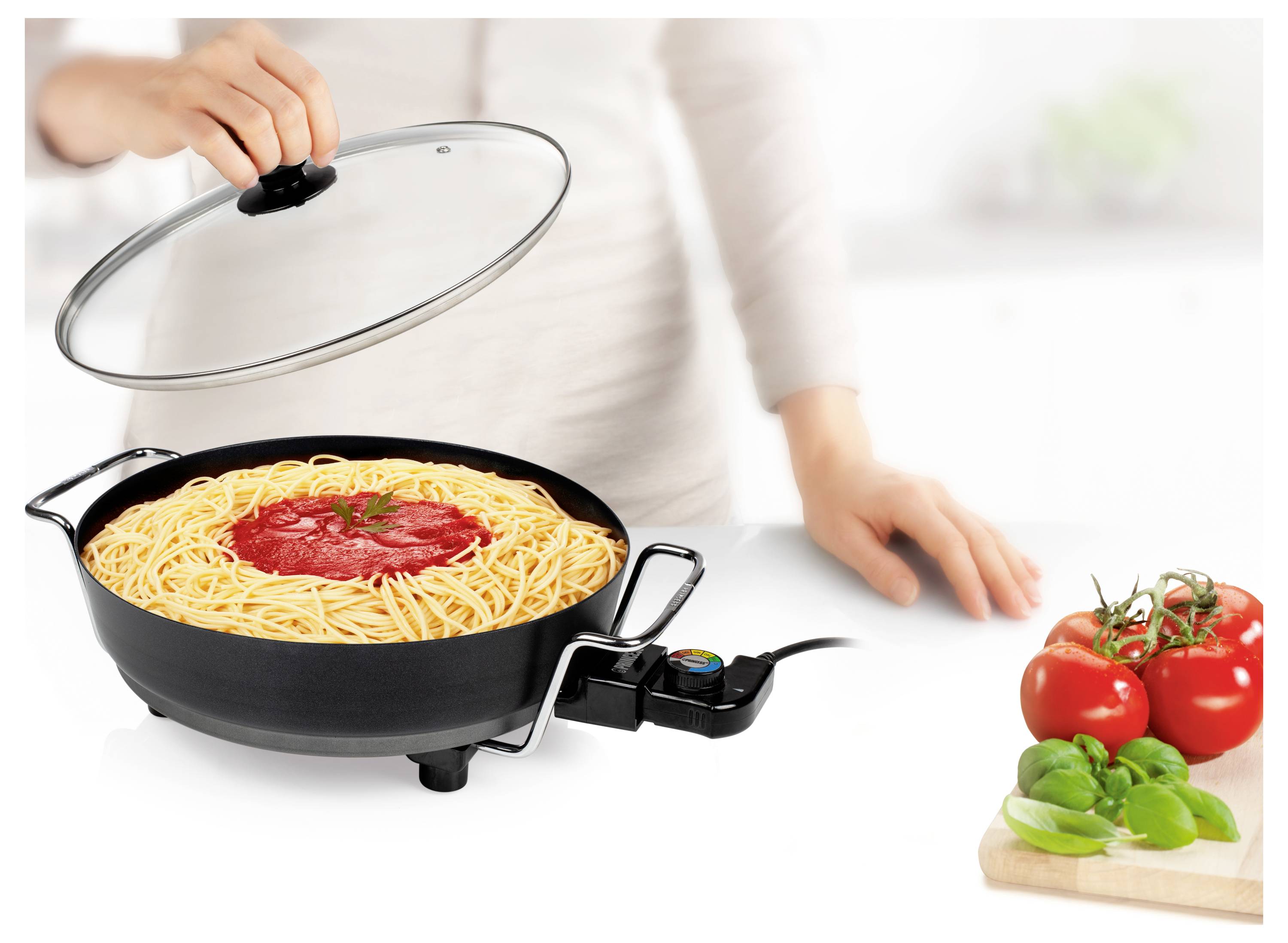 En person lyfter på ett lock och avslöjar spagetti med tomatsås som tillagas i en elektrisk stekpanna bredvid färska tomater och basilika på en vit bänk.